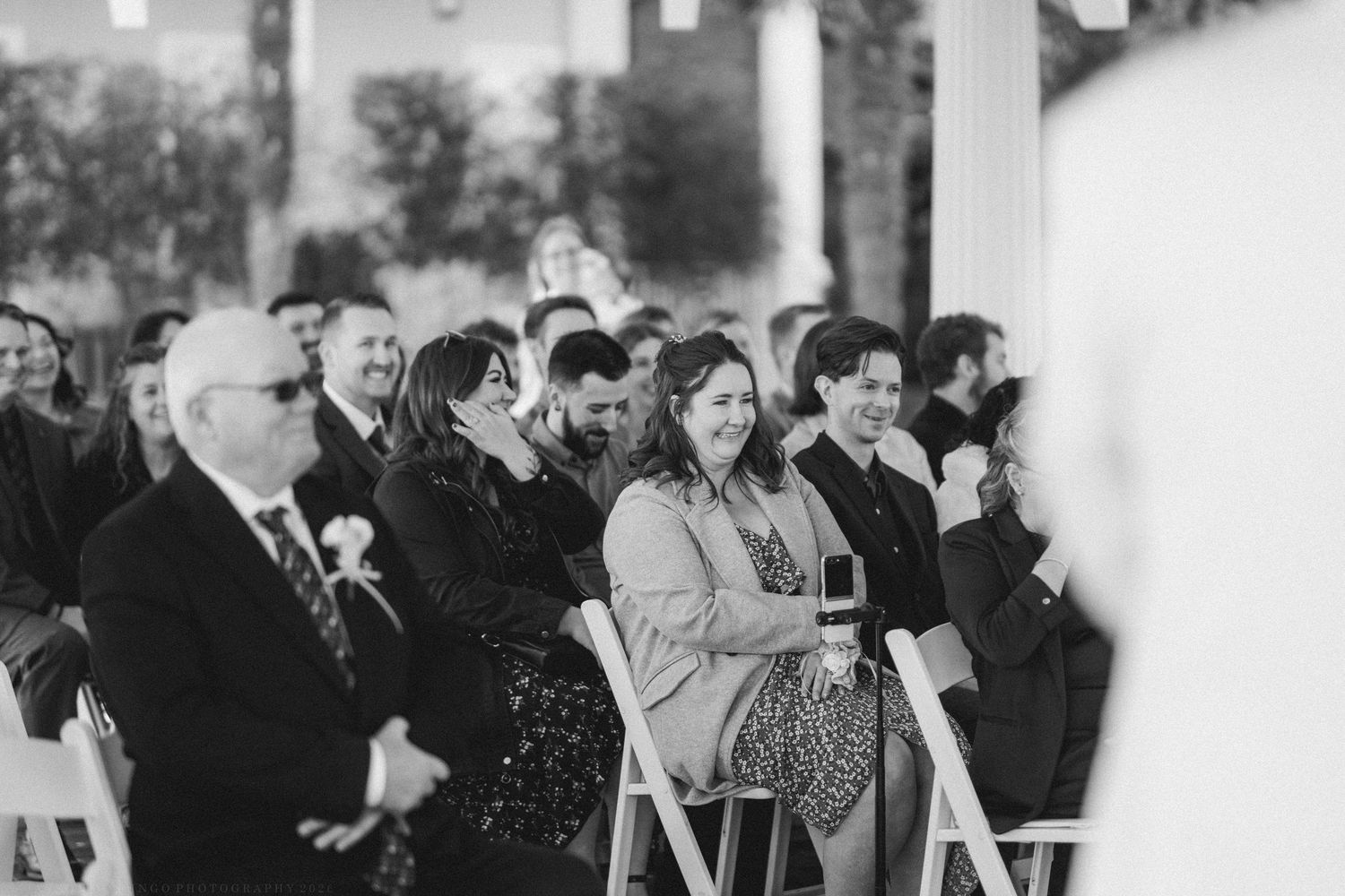 Wedding guests smiling and laughing while seated in white chairs during an outdoor ceremony.