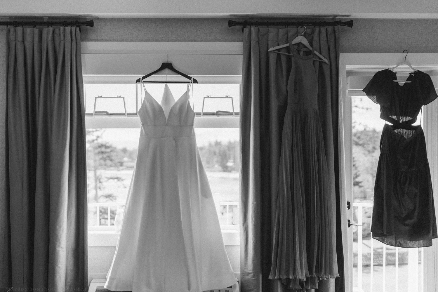 Black and white photo of a wedding dress and bridesmaid gowns hanging by a window before the ceremony.