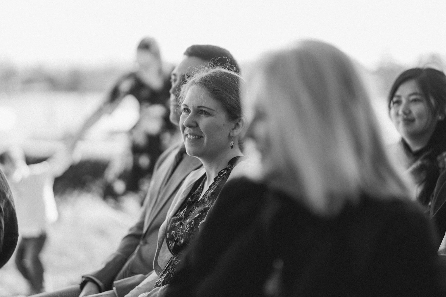 A woman smiles warmly while seated at an outdoor wedding ceremony in black and white photography.