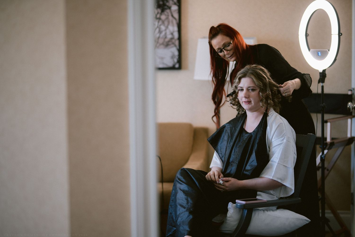 Bride getting her hair styled by a redheaded stylist in a salon chair with a ring light nearby.