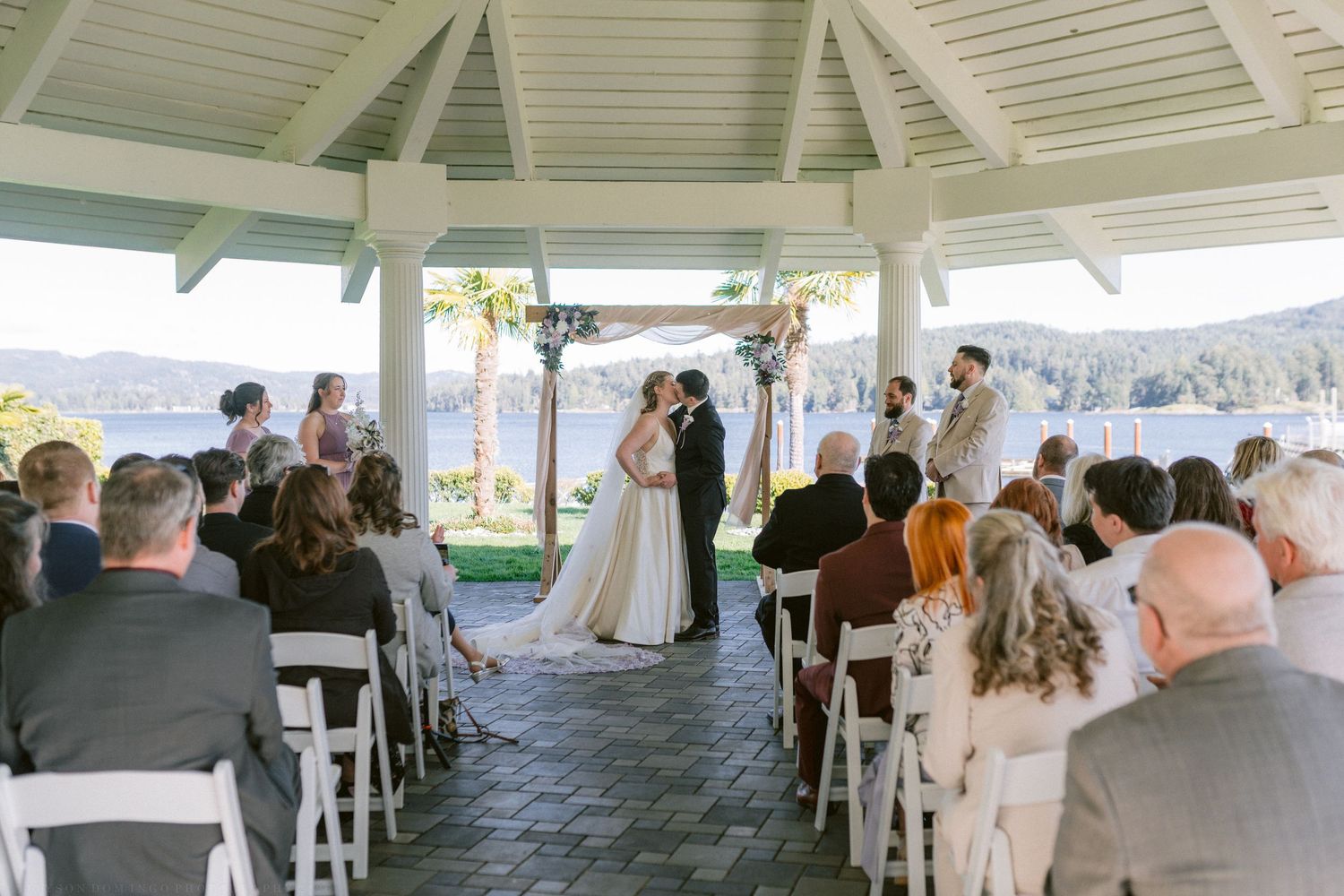Romantic outdoor wedding ceremony under a white gazebo overlooking a serene lake with guests seated in white chairs.