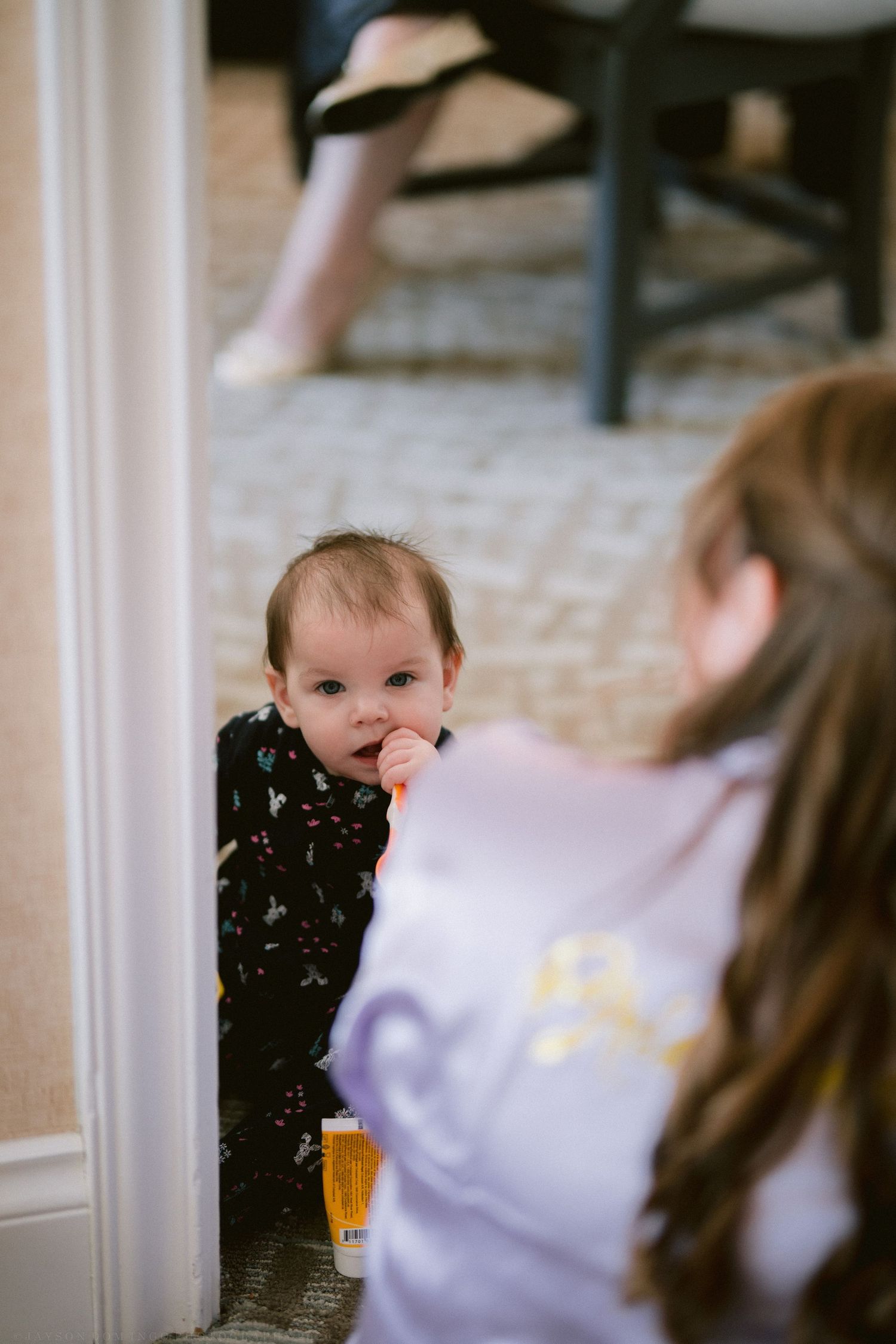 Curious baby in floral outfit looks up at bridesmaid in lavender robe during wedding preparations.