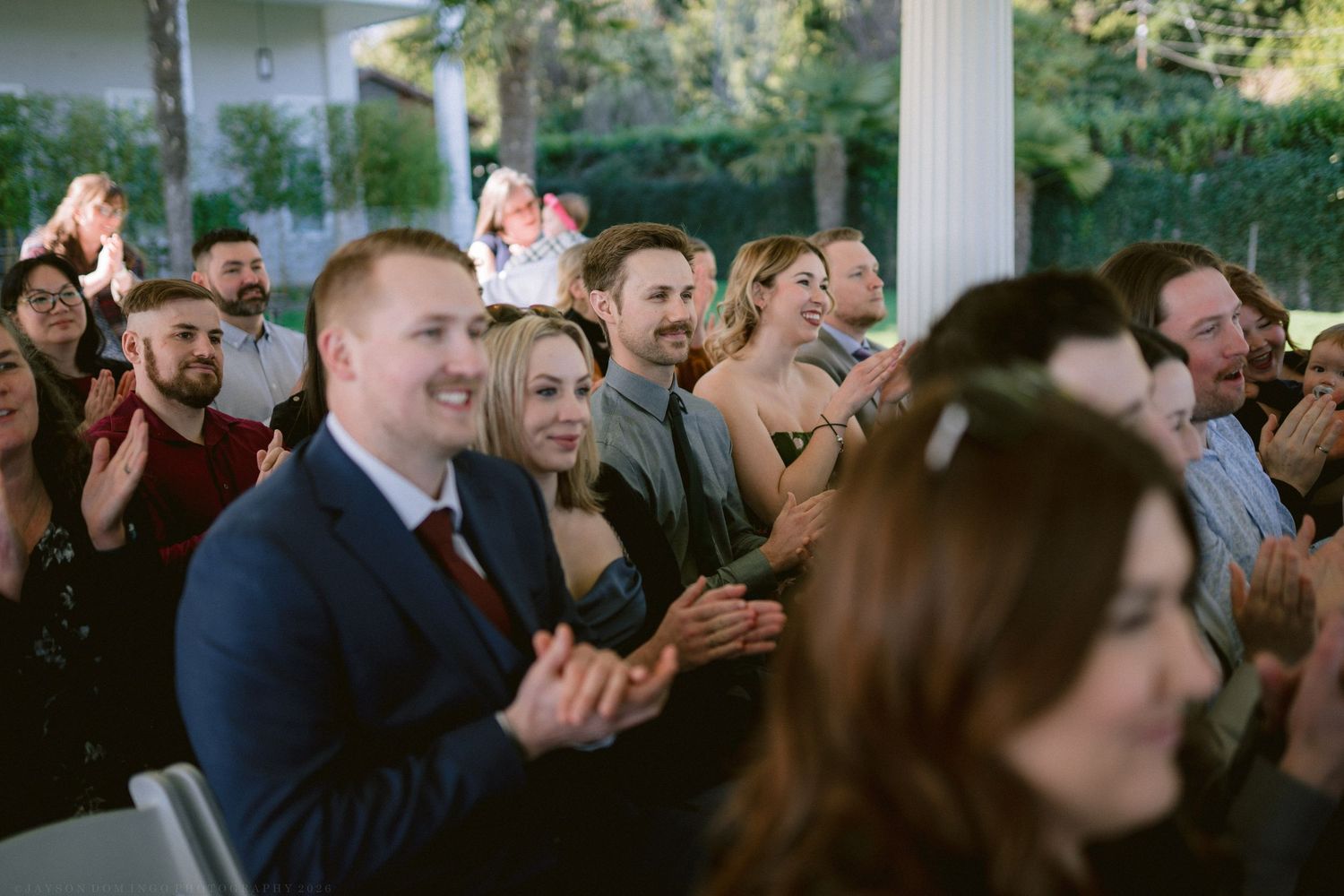 Wedding guests smiling and clapping during a ceremony at an elegant outdoor venue with white columns.