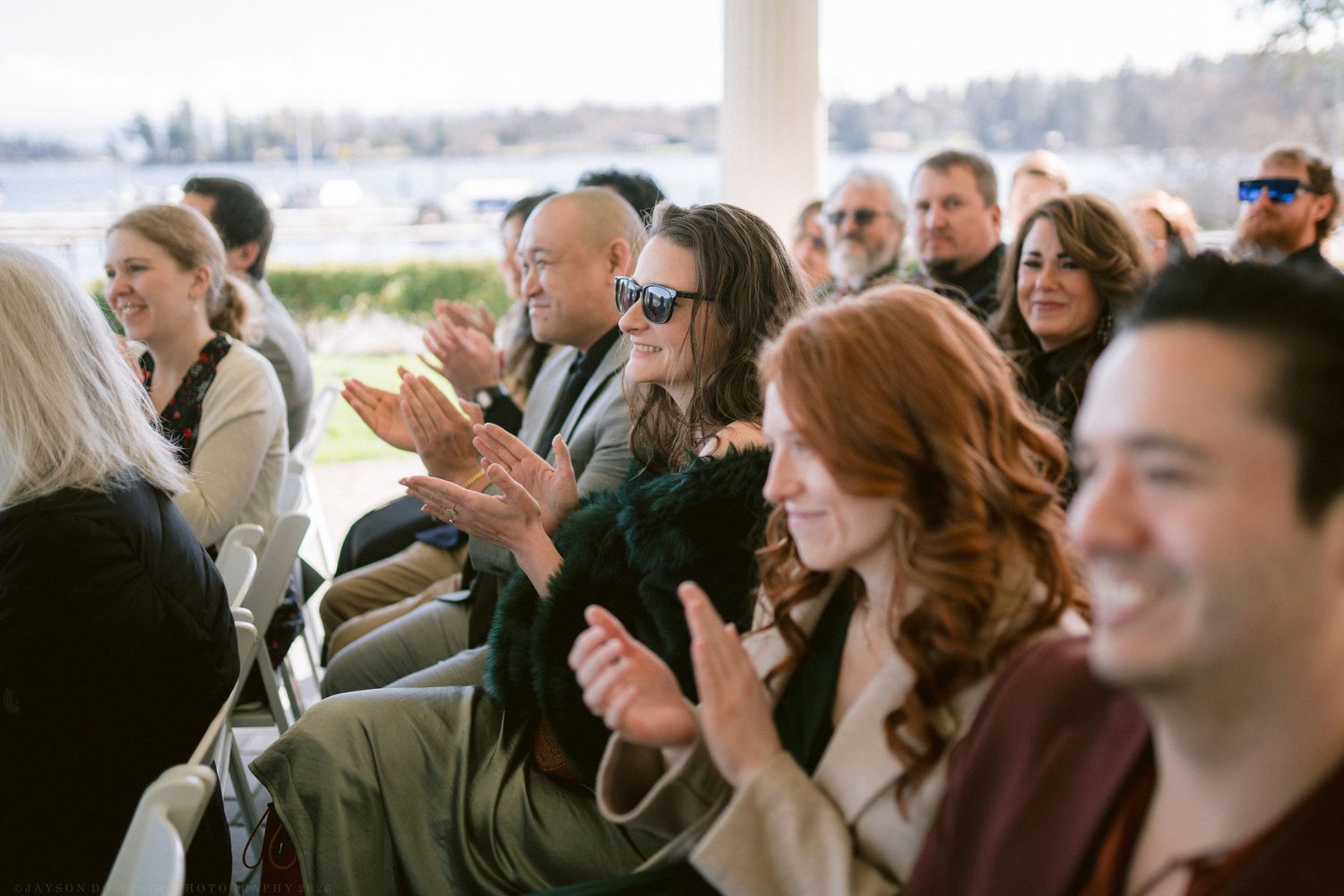 Wedding guests clapping and smiling during an outdoor ceremony by the waterfront.