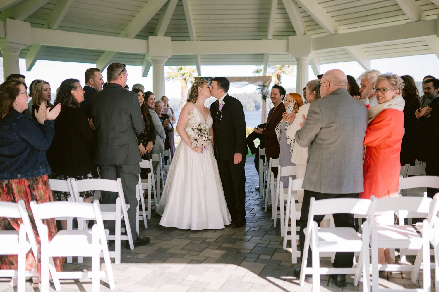 Bride and groom share first kiss at gazebo wedding ceremony with guests applauding.