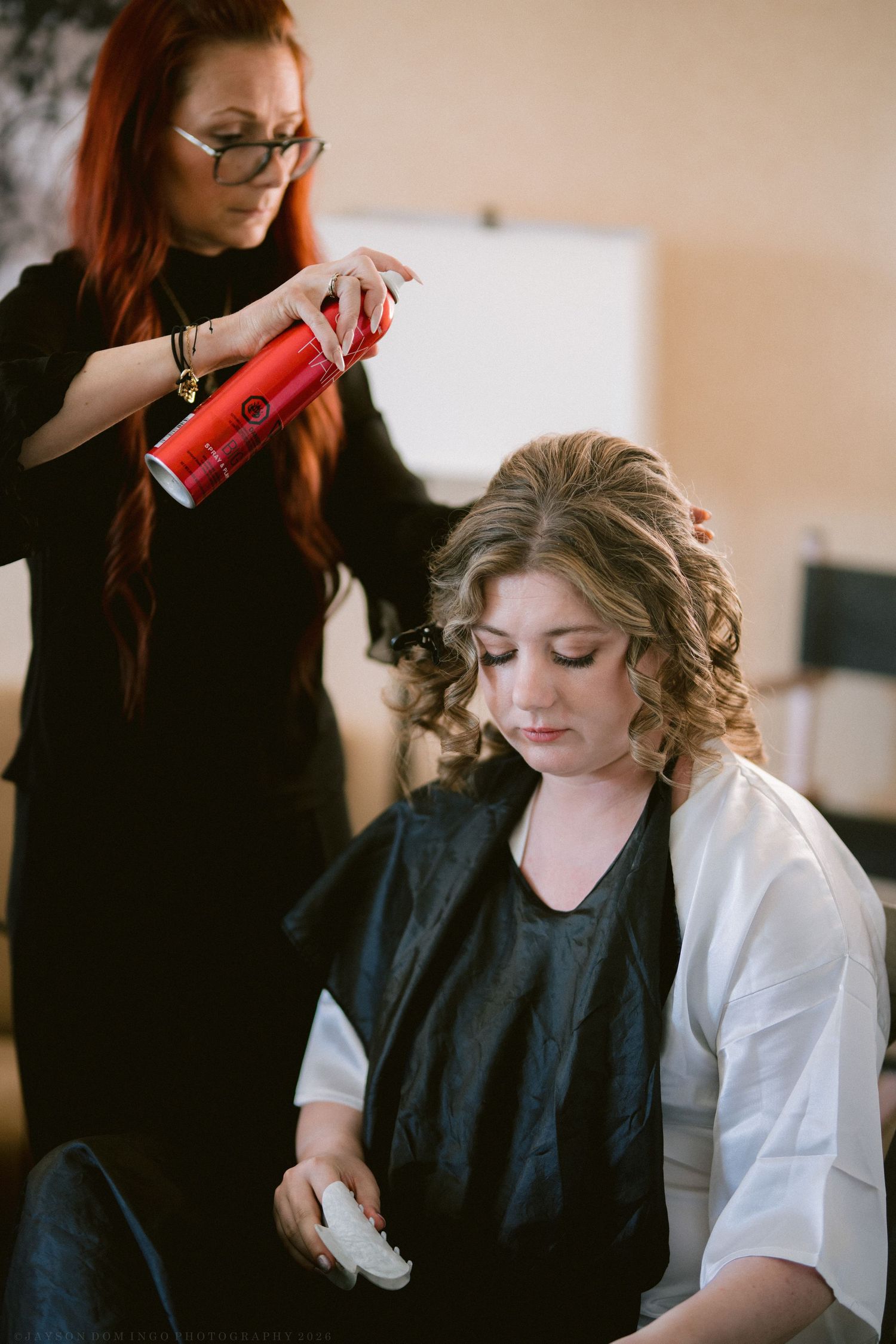 Red-haired stylist applying hairspray to bride's curly updo during wedding day preparations.