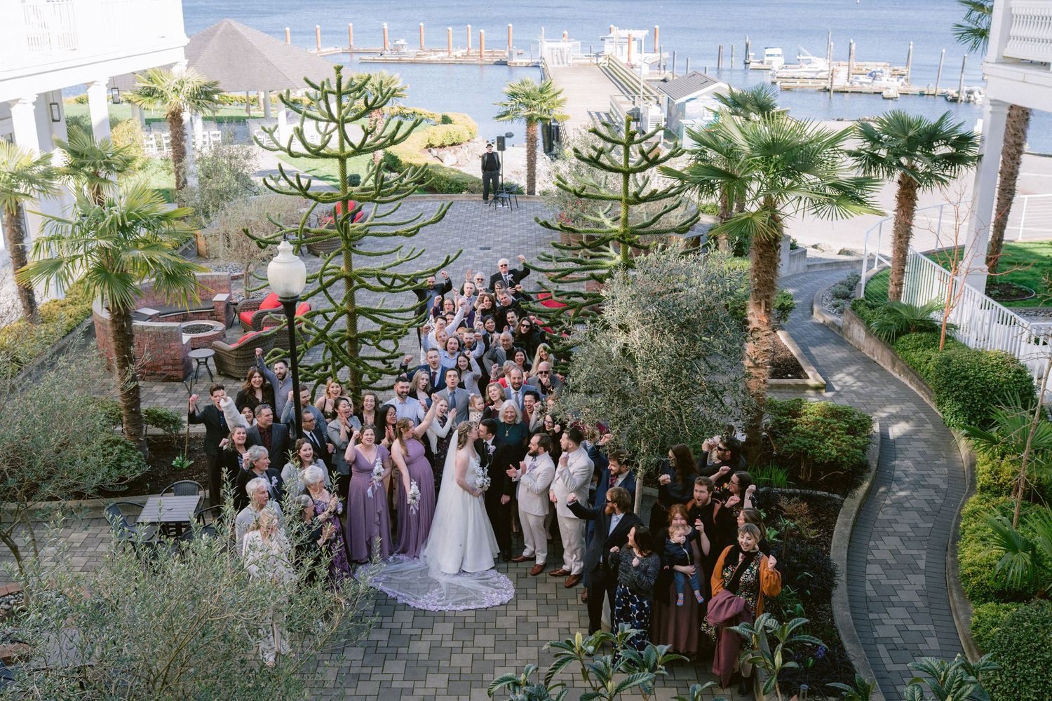 Aerial view of wedding guests gathered in a waterfront garden courtyard with palm trees during an outdoor ceremony.