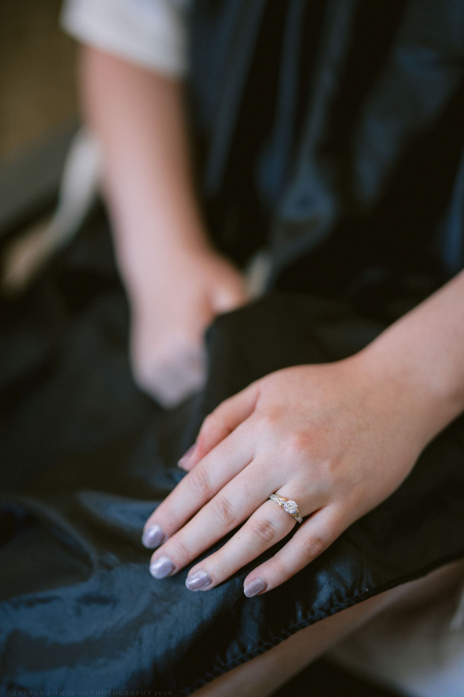 Close-up of a bride's hand displaying a delicate diamond engagement ring with soft lavender nail polish on dark fabric.