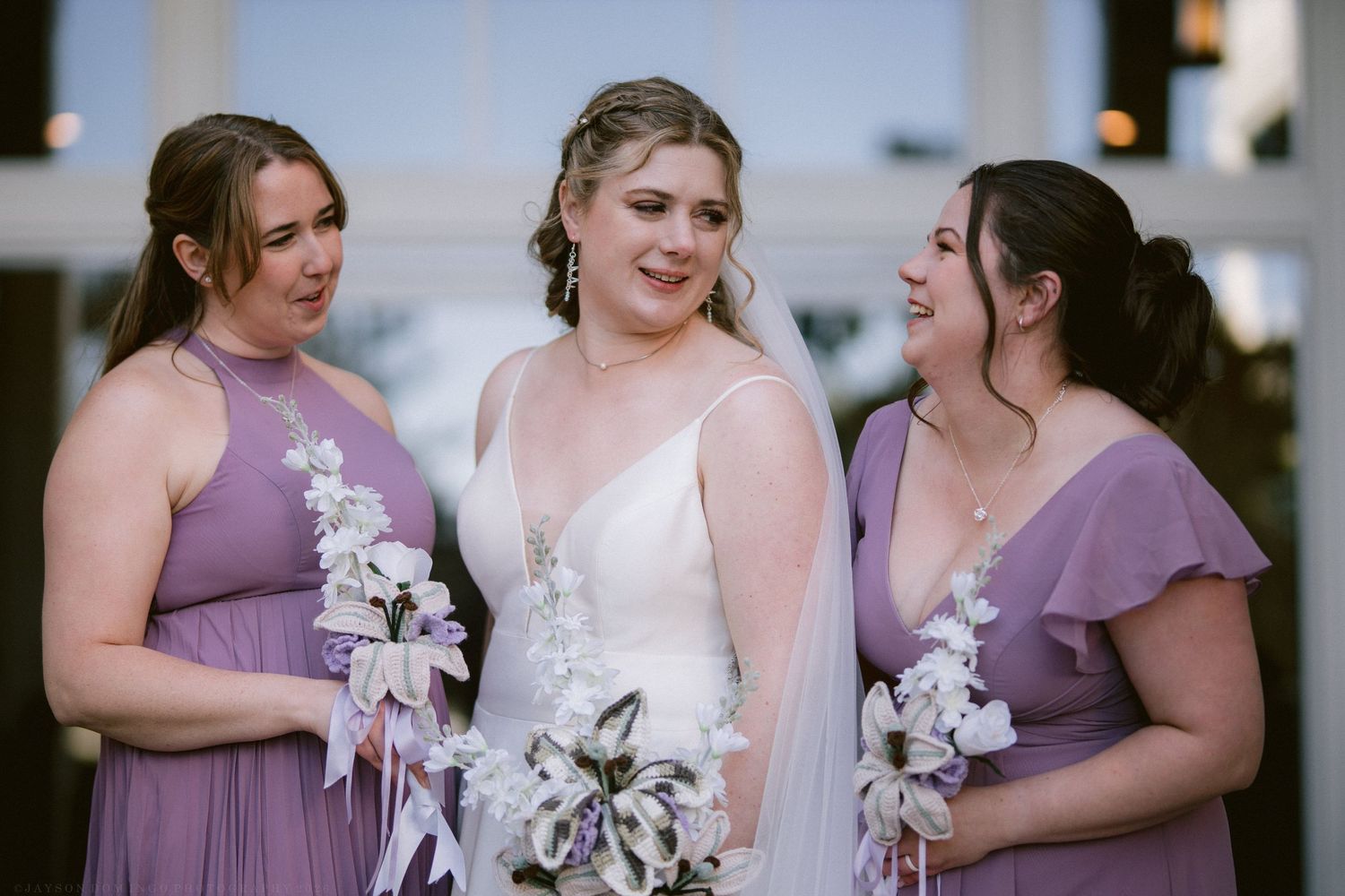 Bride in white gown and veil stands smiling with two bridesmaids in purple dresses holding bouquets.