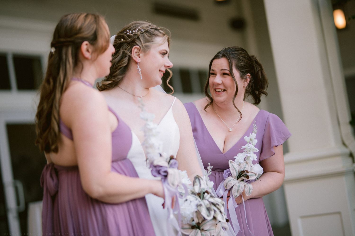 Bride and bridesmaids in lavender dresses laughing together while holding floral bouquets outdoors.