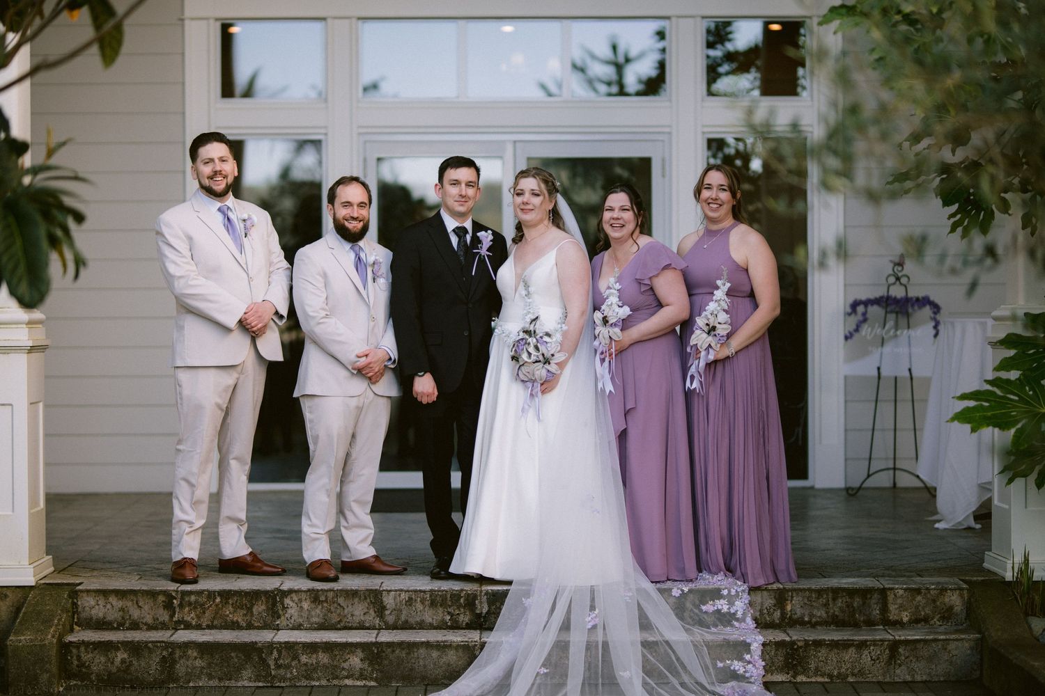 Wedding party poses on steps featuring bride in white gown with bridesmaids in purple dresses and groomsmen.
