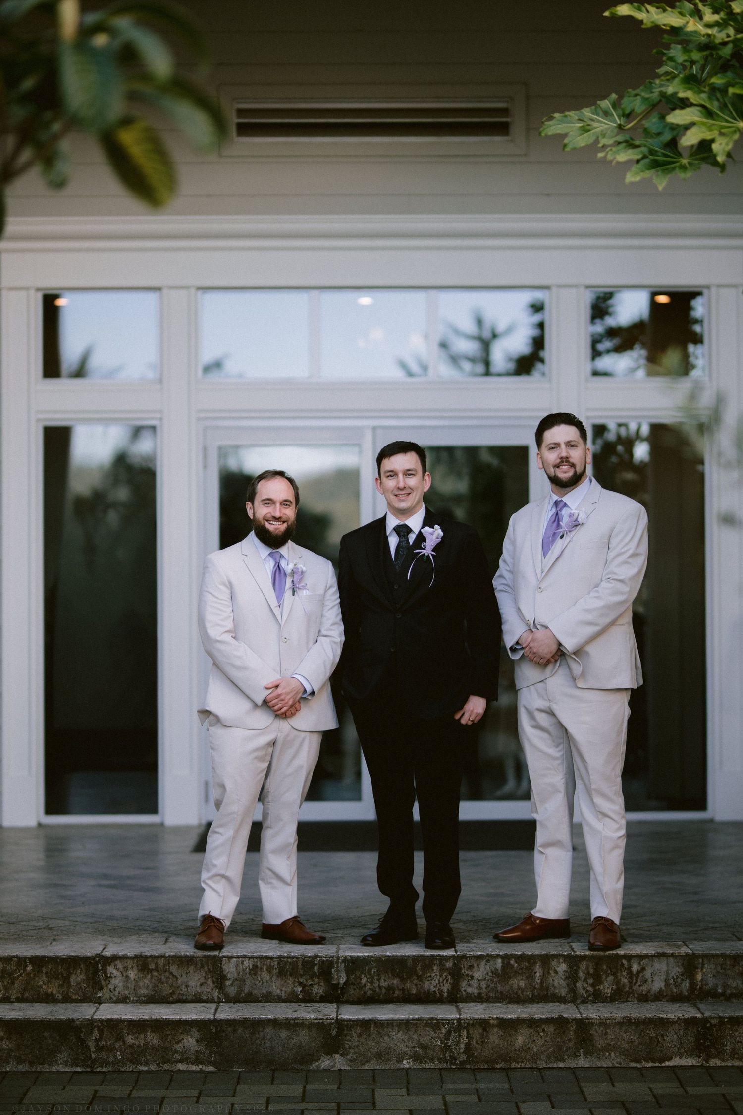 Groom in black suit stands with two groomsmen in tan suits and lavender ties outdoors.