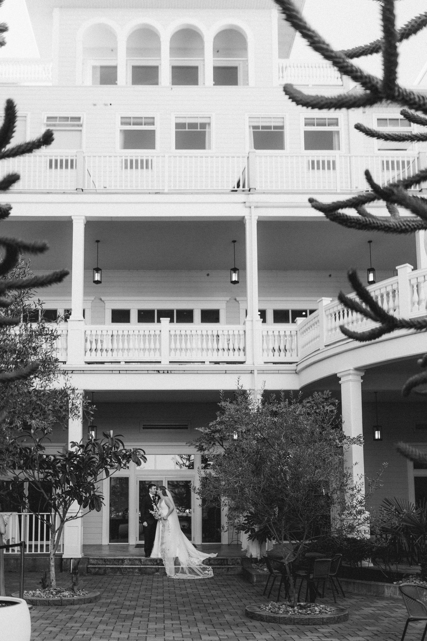 Black and white photo of bride and groom embracing outside elegant multi-story venue.