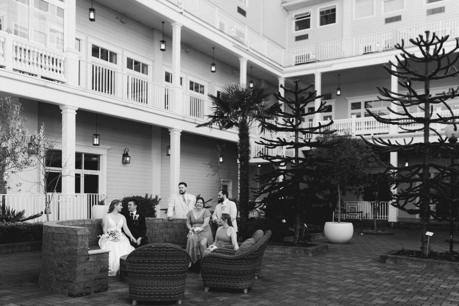 Black and white photo of wedding party relaxing on patio furniture outside grand hotel.
