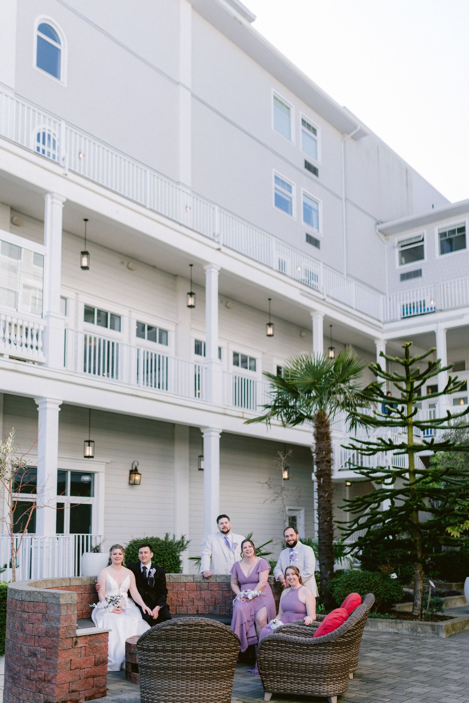 Wedding party poses in wicker chairs on a patio courtyard of a elegant white multi-story hotel venue.