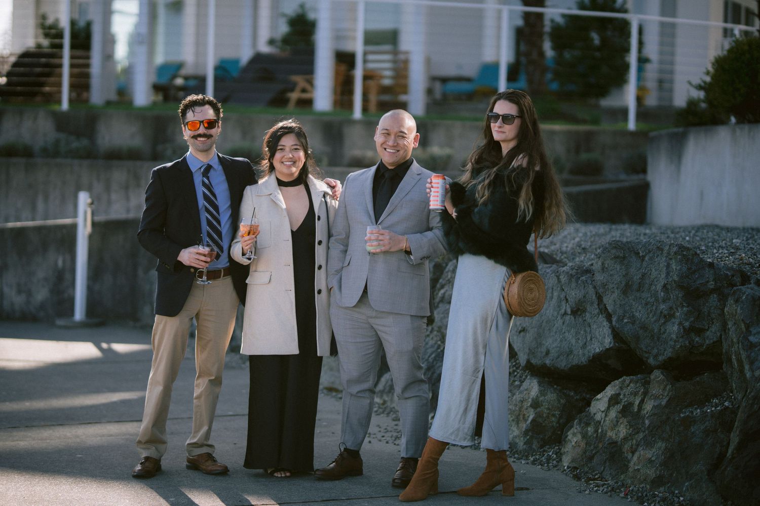 Four elegantly dressed wedding guests posing outdoors near a waterfront venue on a sunny day.