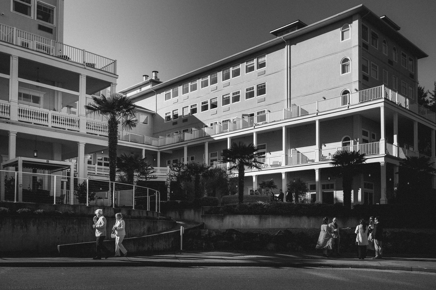 Black and white photo of a multi-story beachfront hotel with palm trees and pedestrians walking along the street.