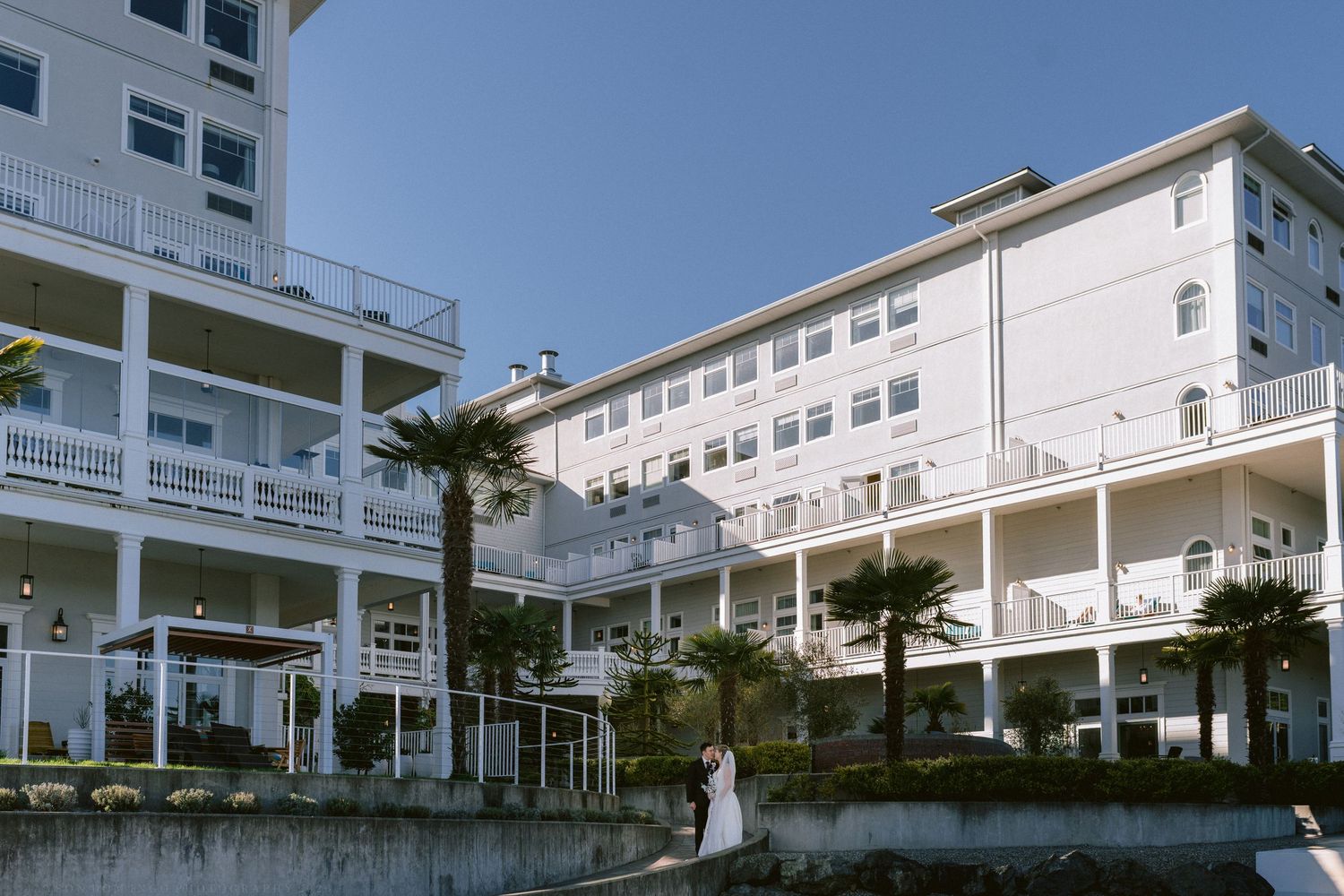 Wedding couple posing outside grand white coastal hotel with palm trees.