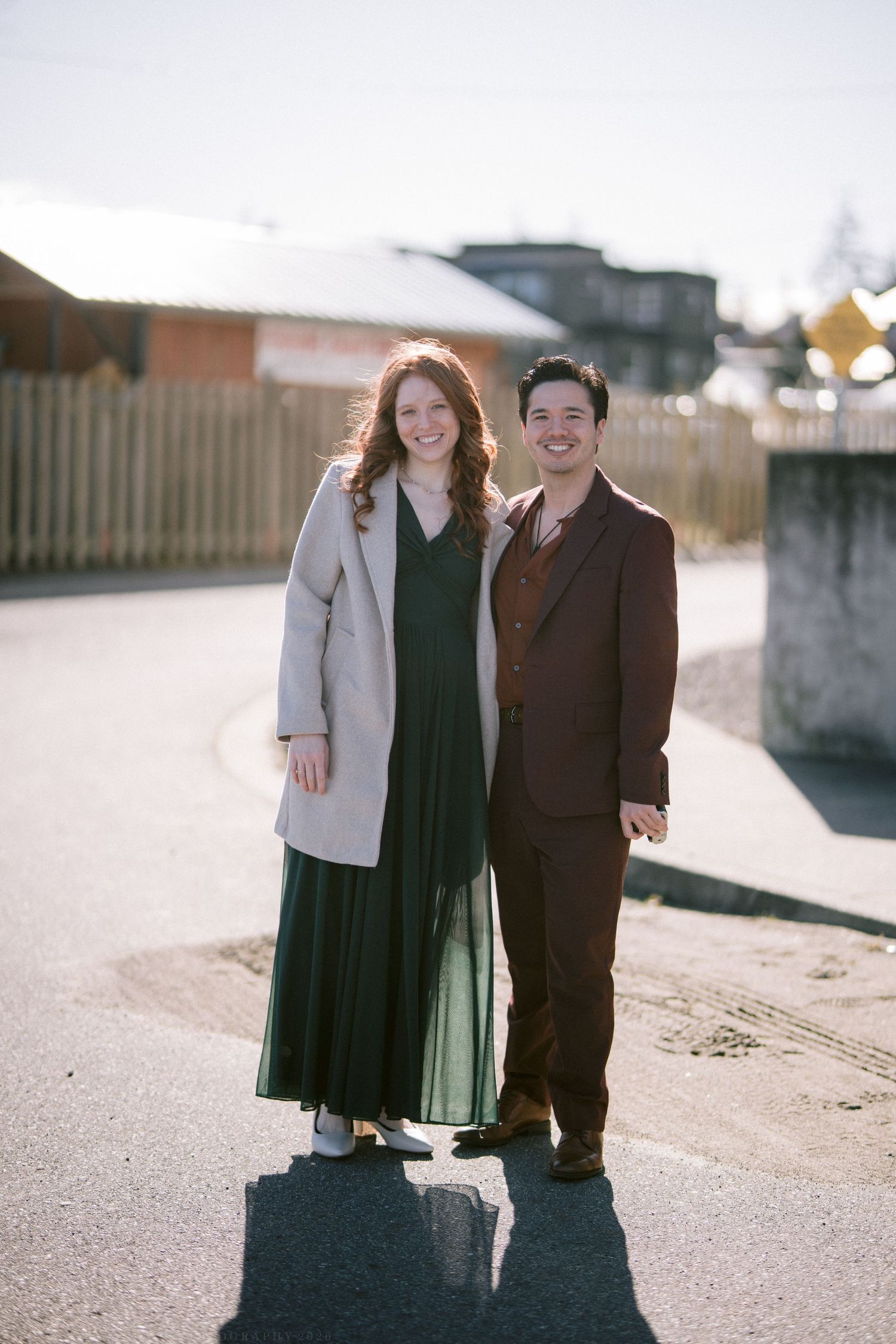 A smiling couple poses outdoors in elegant wedding guest attire on a sunny day.