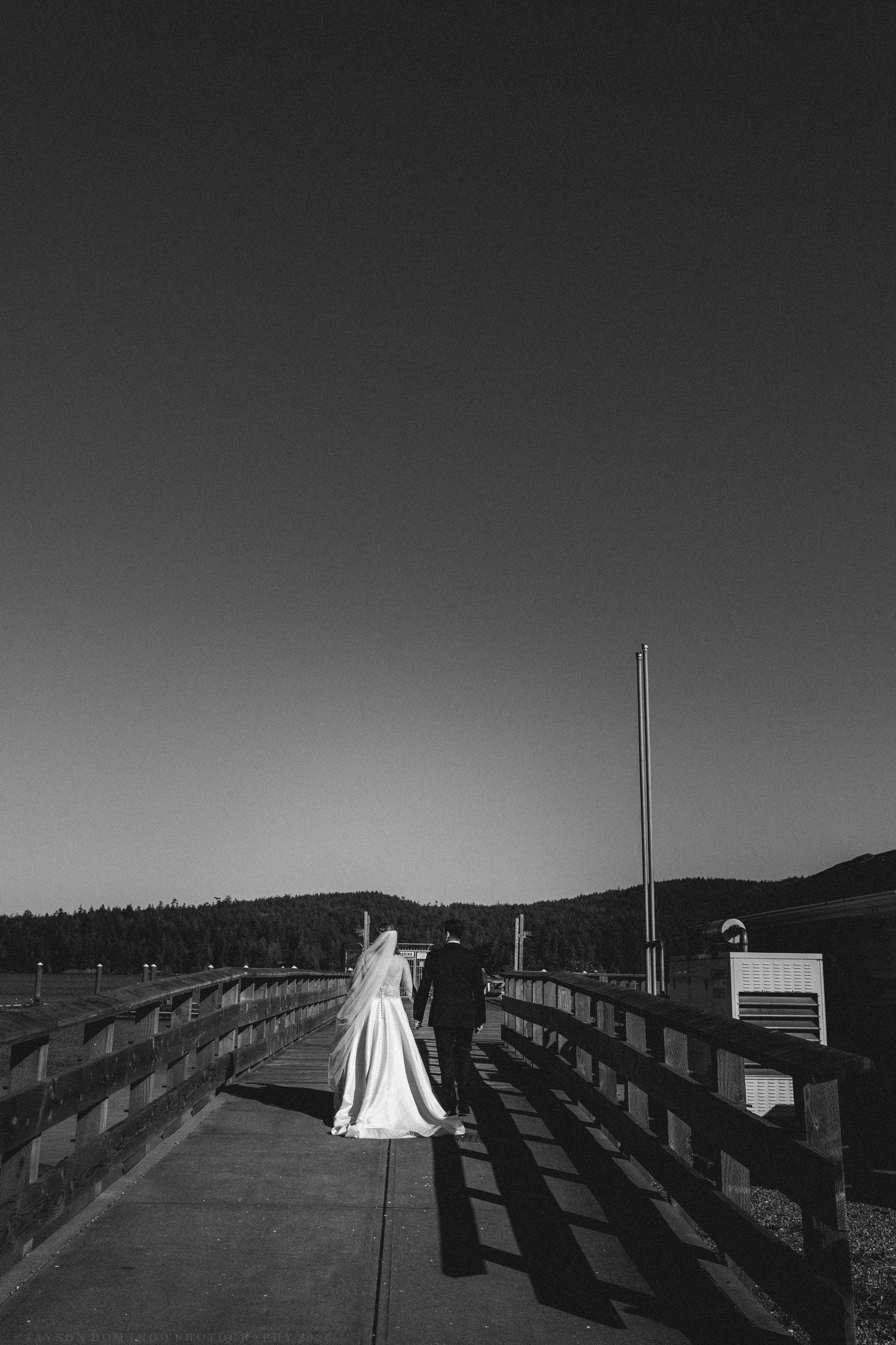Black and white photo of bride and groom walking along a wooden pier under a clear sky.