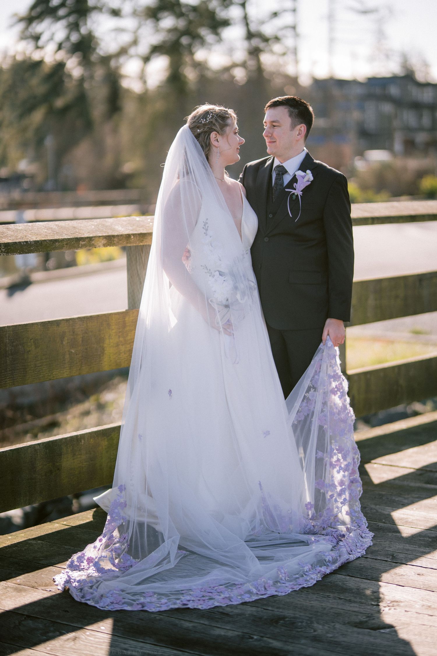 Bride and groom sharing a romantic moment on a wooden bridge, bride wearing a long veil with lace trim.