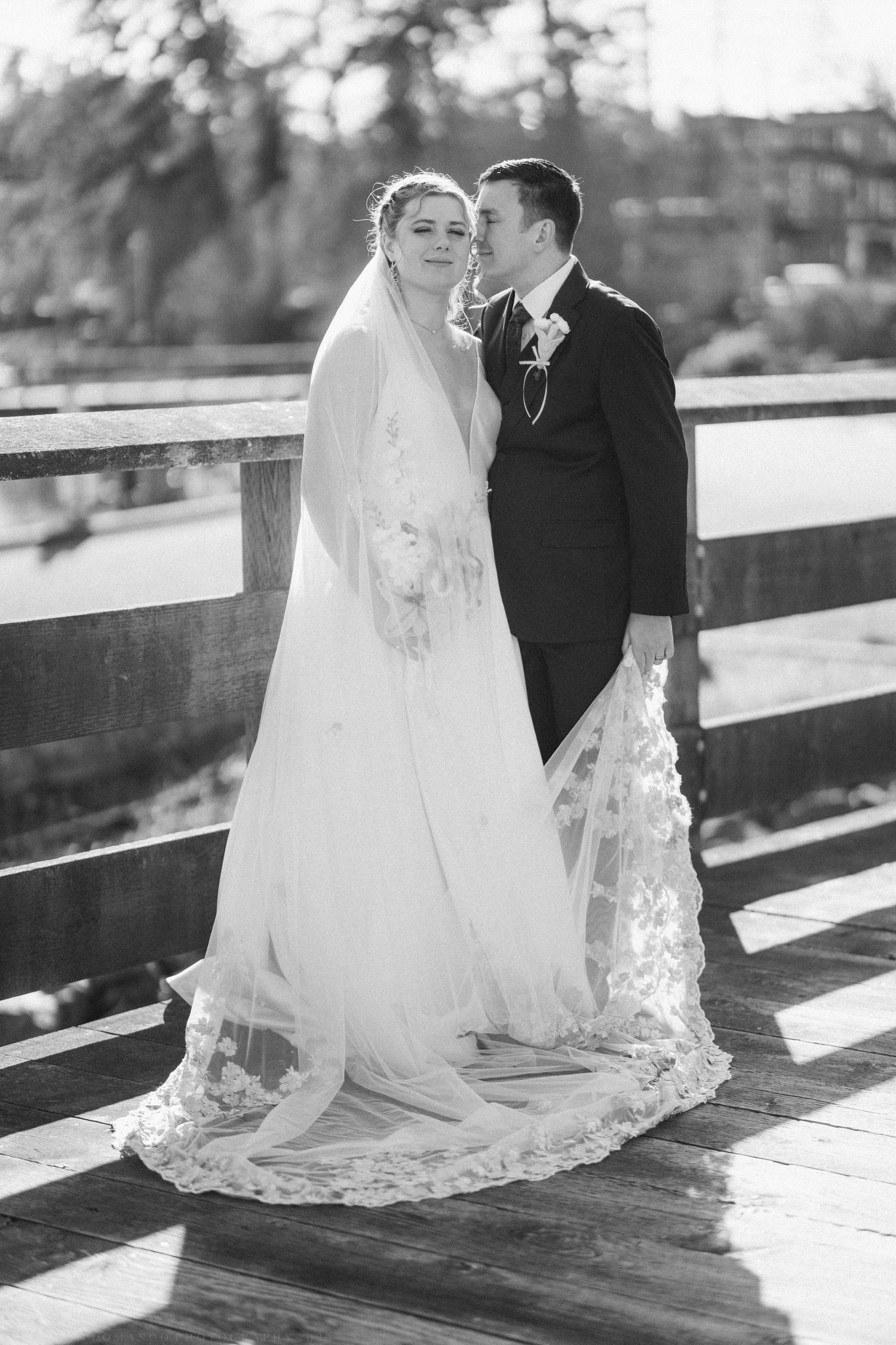 Black and white photo of a bride and groom sharing a tender moment on a wooden bridge, her lace veil trailing behind.