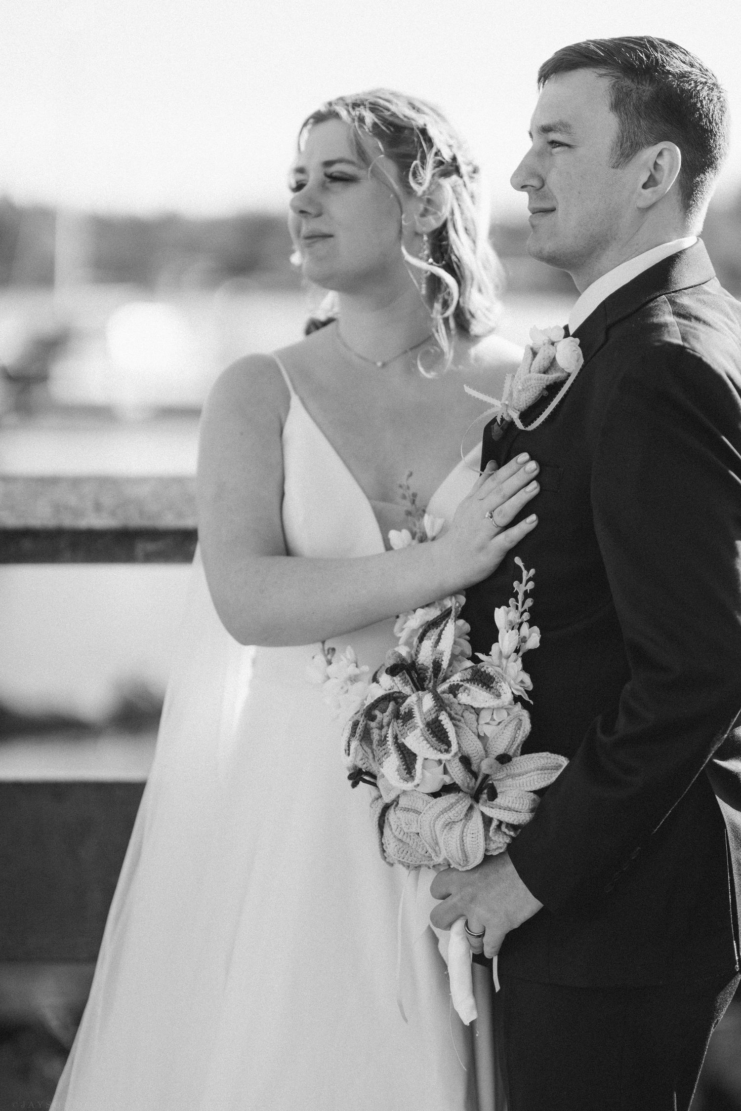 Black and white portrait of bride holding bouquet standing close with groom near waterfront.