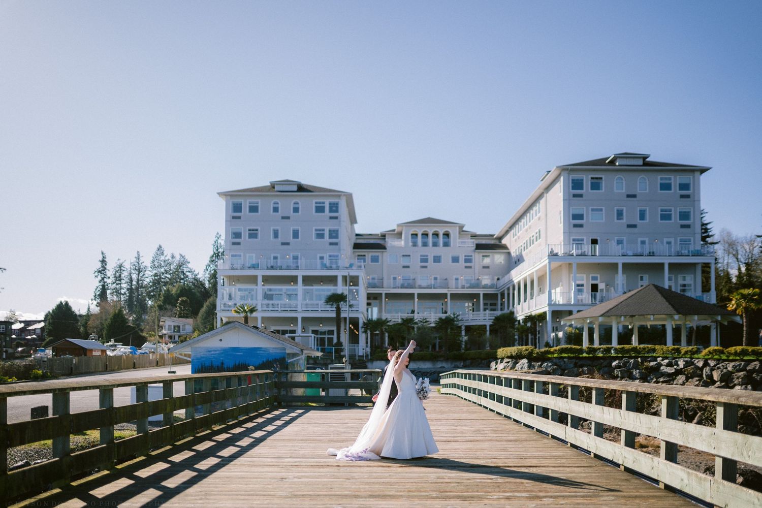 Bride in flowing white gown stands on wooden dock before a grand white waterfront hotel on a sunny day.