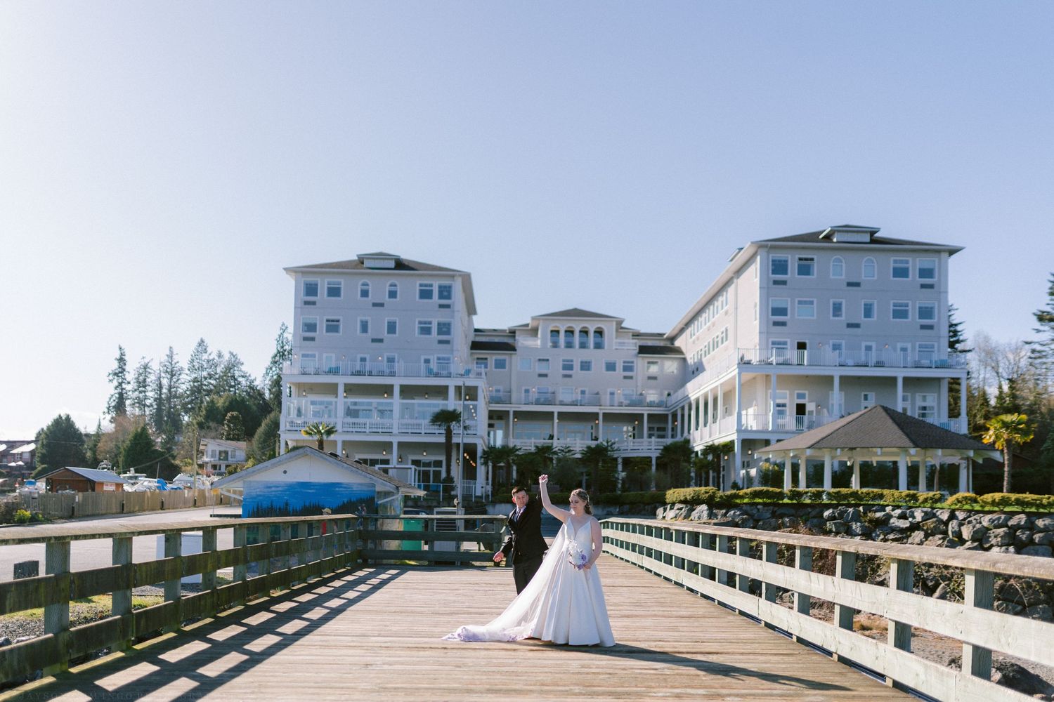 Bride and groom share a romantic moment on a wooden pier with a grand white waterfront hotel behind them.