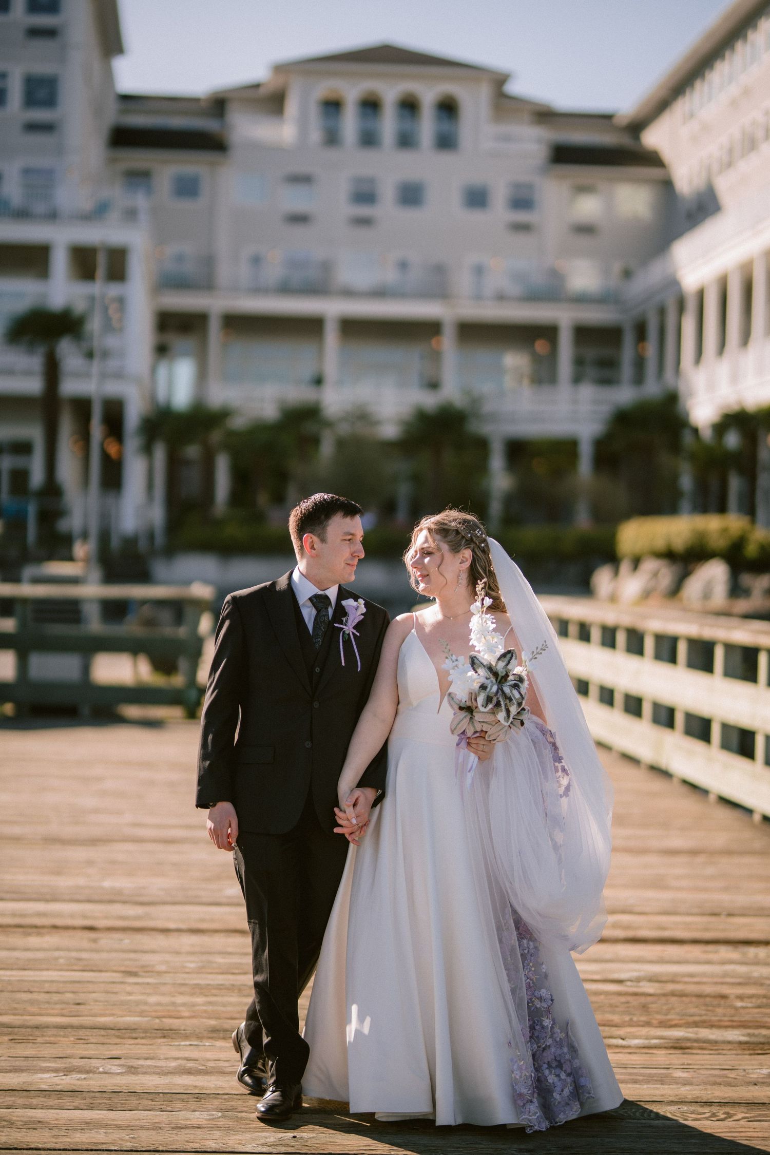 Bride and groom holding hands on a sunny boardwalk with a grand white hotel in the background.