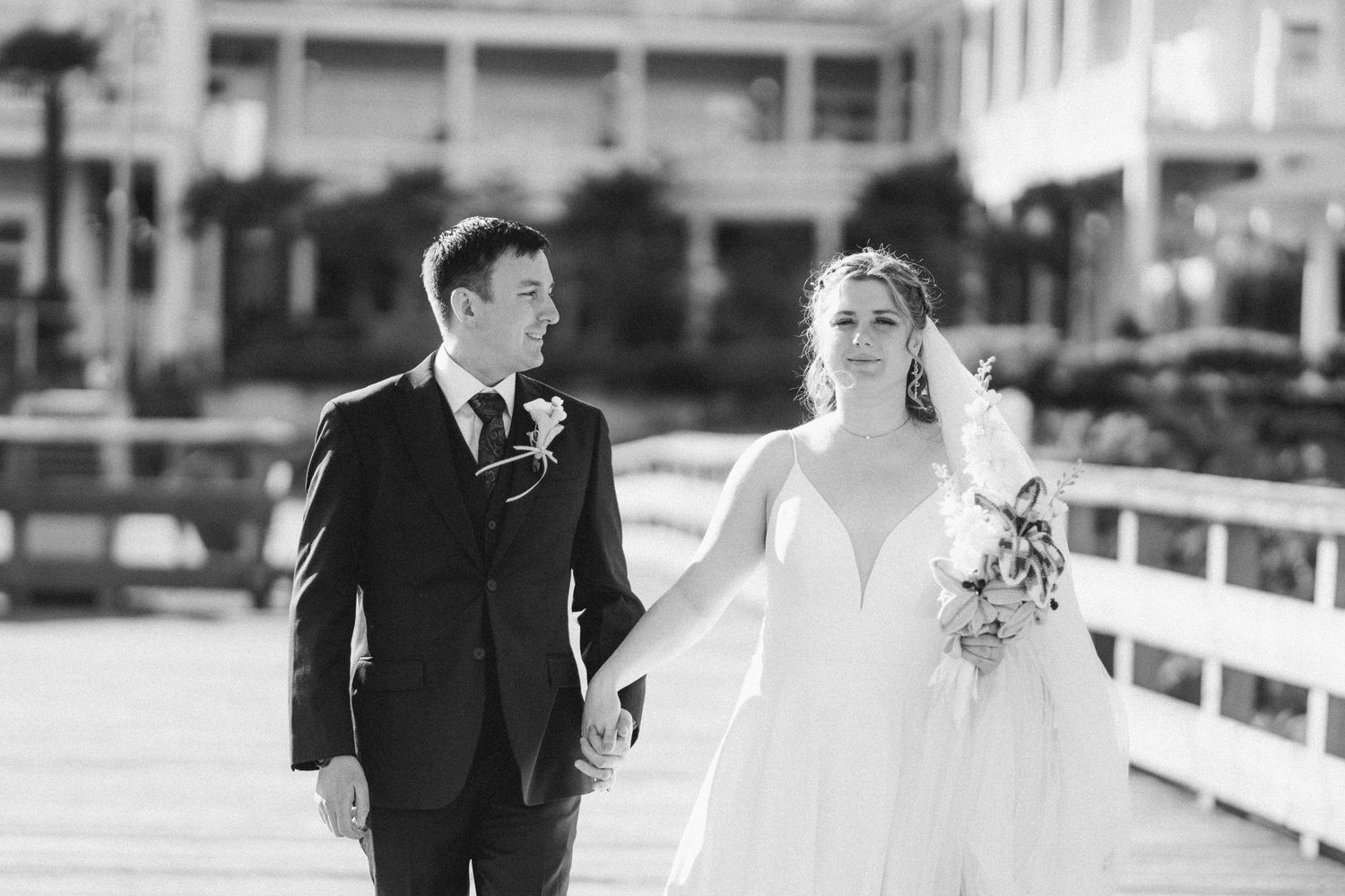 Black and white photo of a bride and groom holding hands while walking along a waterfront pier.