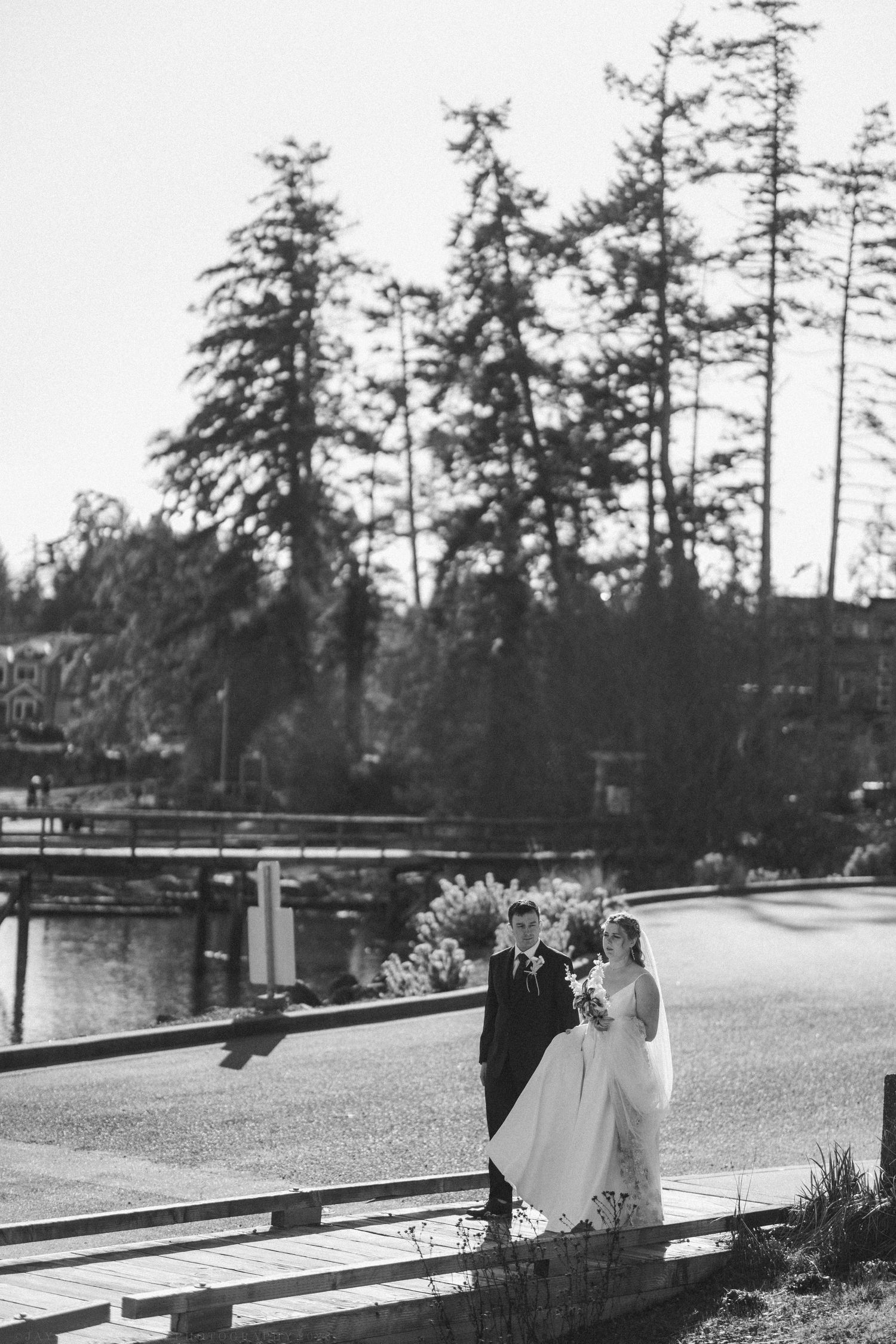Black and white photo of bride and groom standing on a dock near a lake surrounded by tall evergreen trees.