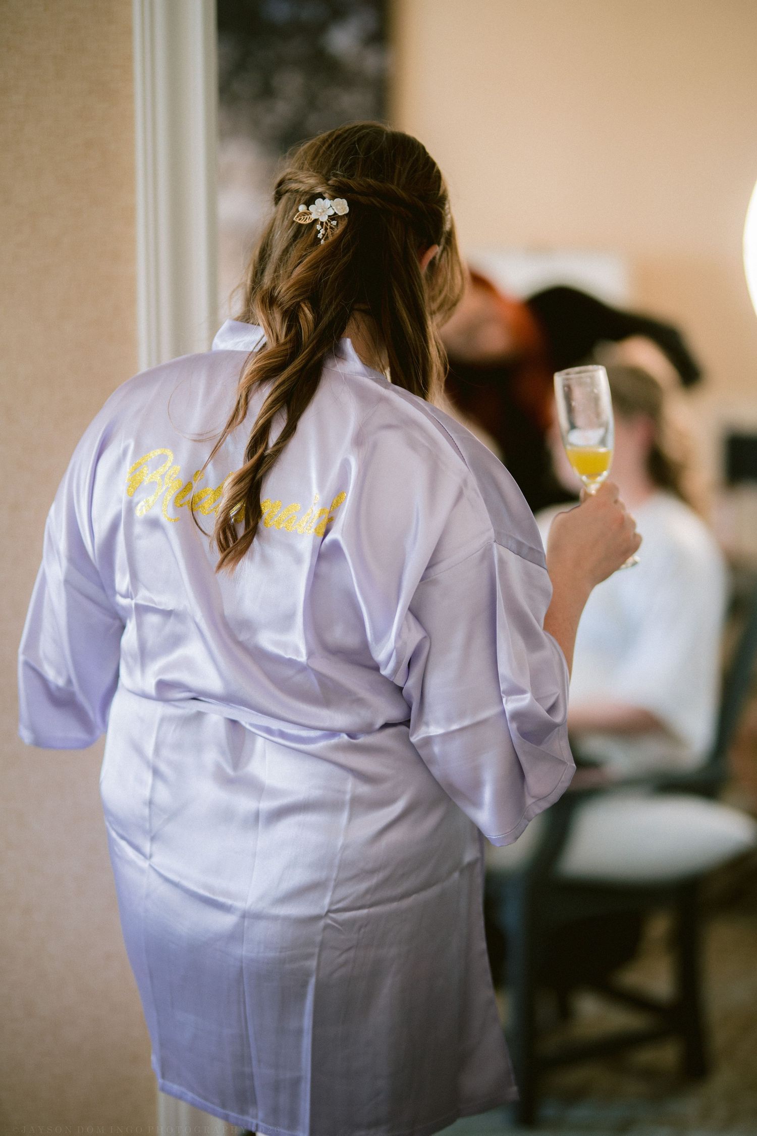 Bridesmaid in lavender satin robe holding champagne glass during bridal party wedding preparations.