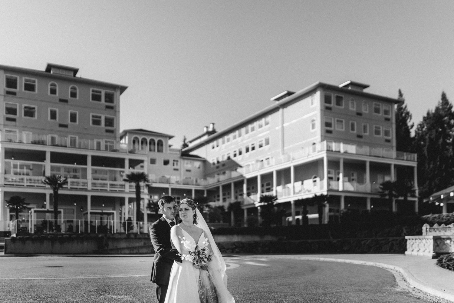 Black and white photo of a bride and groom walking outside a grand multi-story hotel wedding venue.