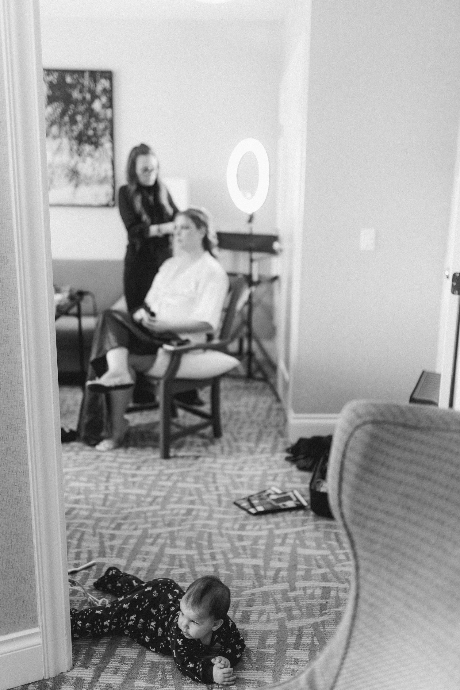 Black and white photo of a bride getting hair done while a baby plays on the floor in a hotel room.