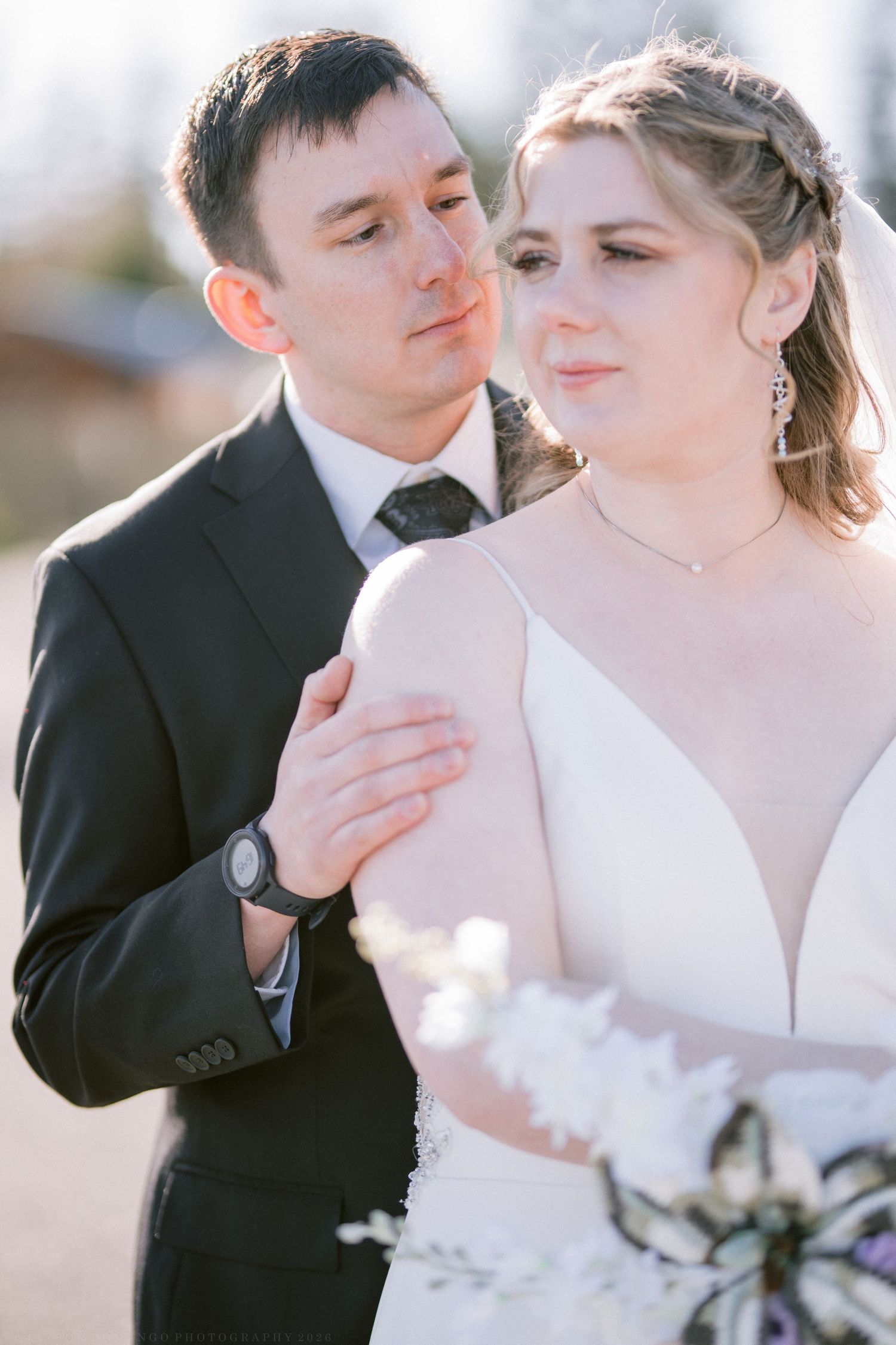 Groom in black suit embraces bride in white wedding gown while she holds a bouquet outdoors.
