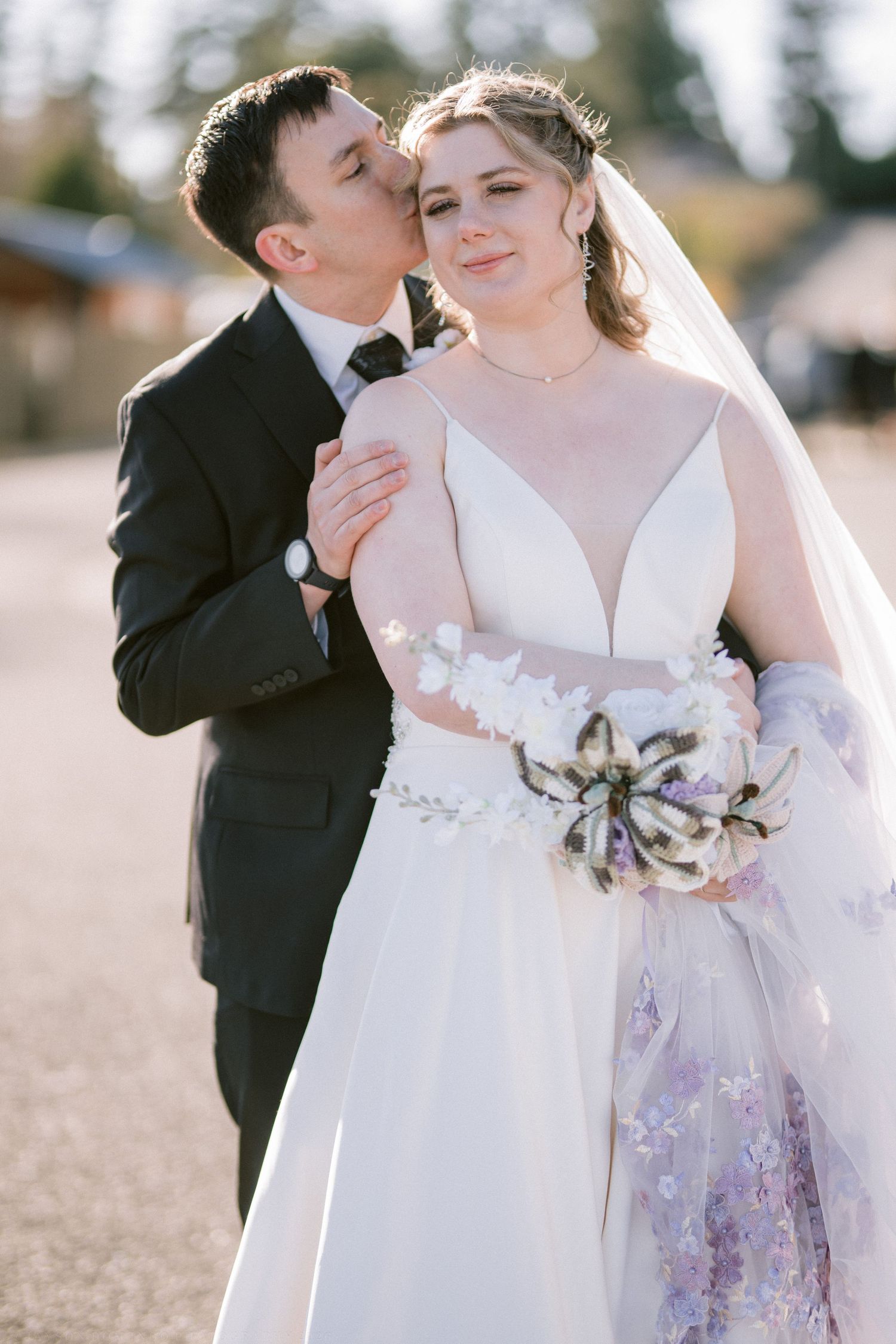 Groom kissing bride on cheek as she holds a floral bouquet in a stunning embroidered wedding gown.