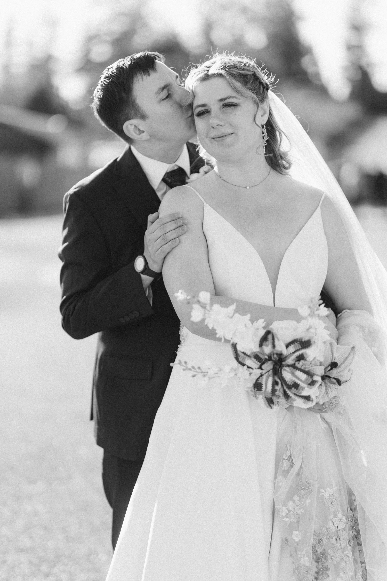 Black and white photo of groom kissing bride on cheek as she smiles holding a wedding bouquet.