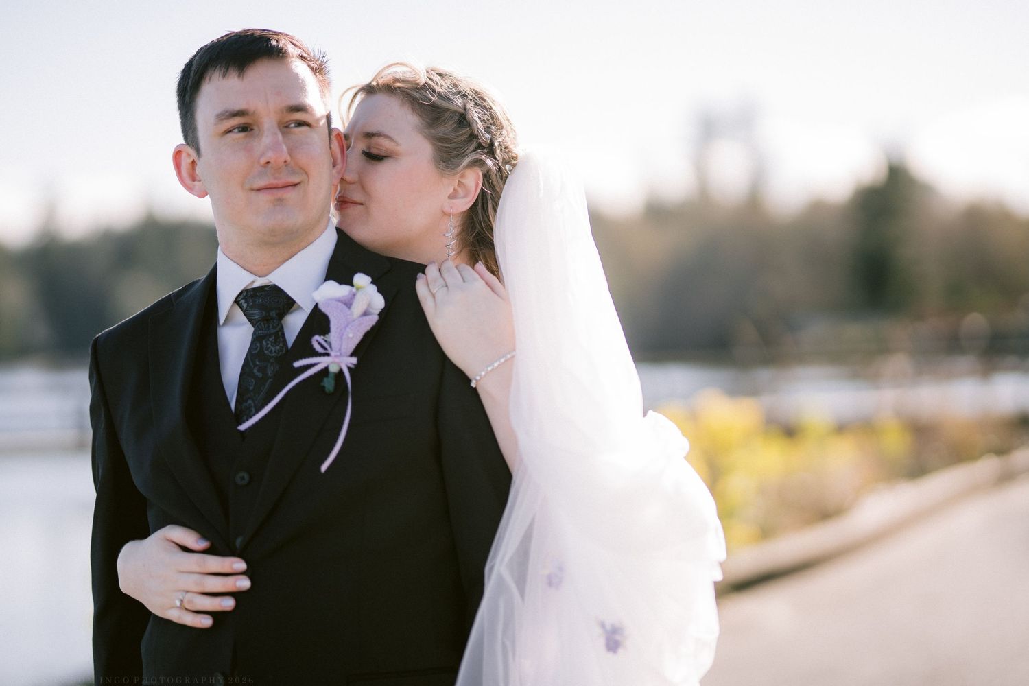 Bride in white veil kisses groom in black suit with purple boutonniere outdoors at wedding.