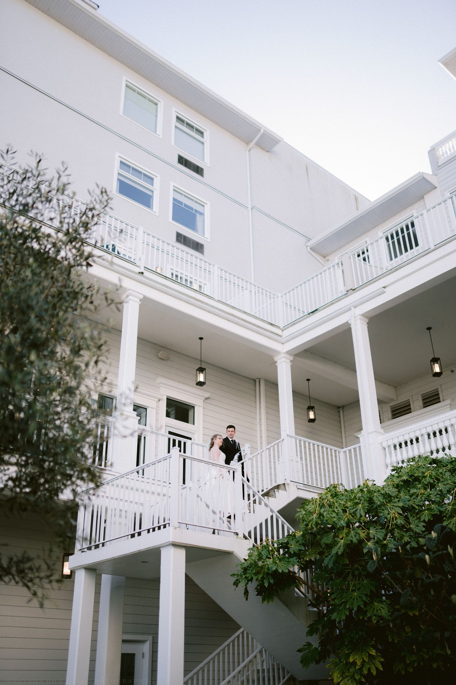 Couple stands on white balcony staircase of elegant multi-story wedding venue building.