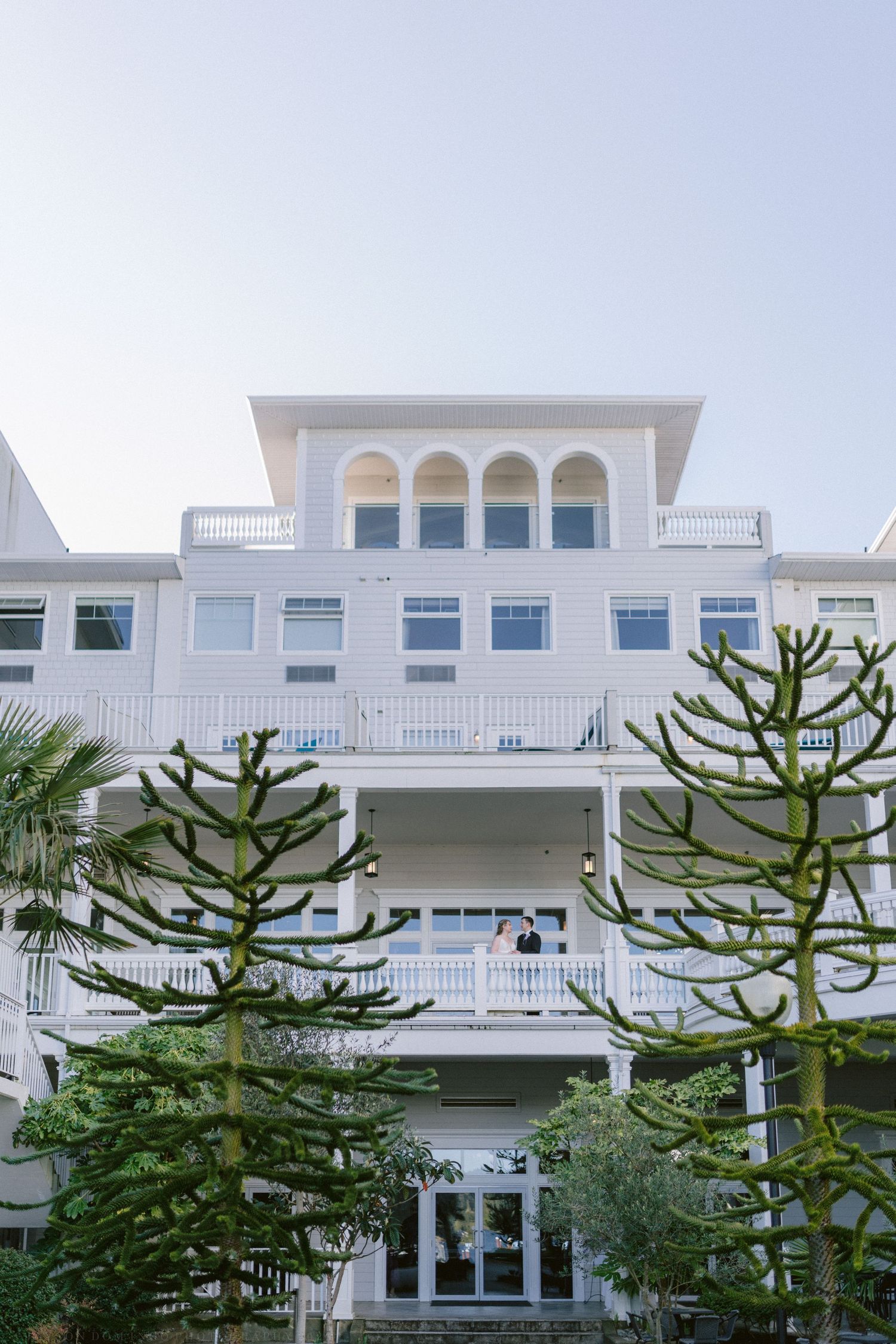 Couple poses on balcony of grand white multi-story coastal wedding venue with tall trees.