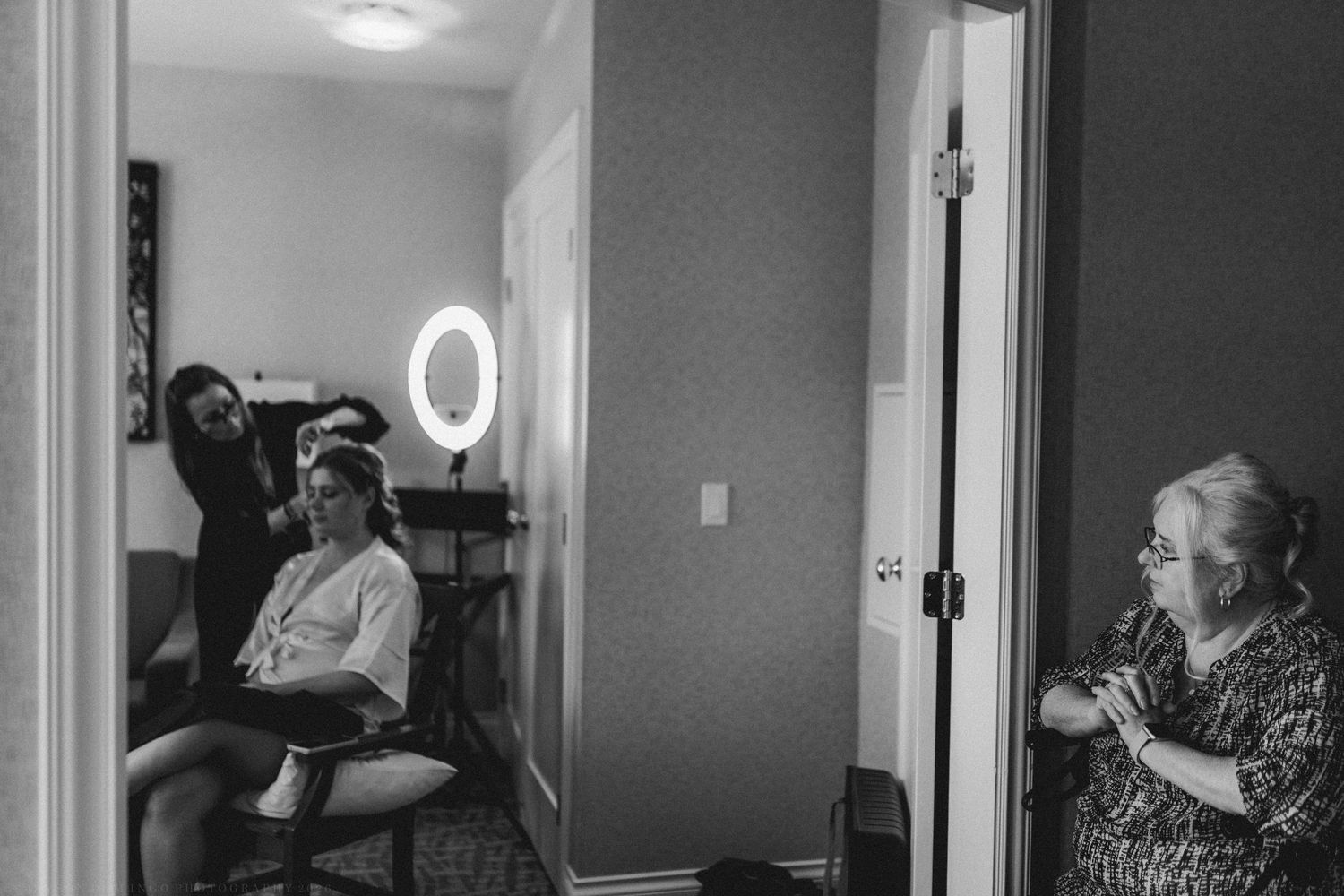 Black and white photo of bride getting hair done while elderly woman watches in doorway.