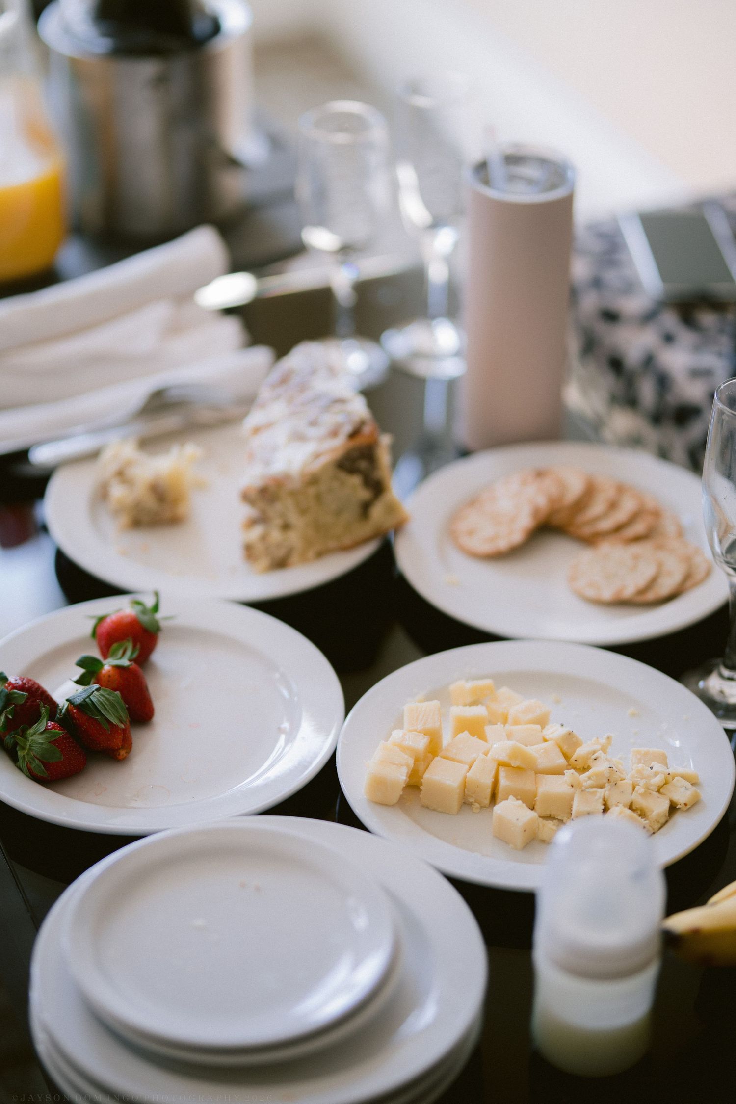 A spread of appetizers including cubed cheese, strawberries, crackers, and cake slices on white plates atop a dark table.