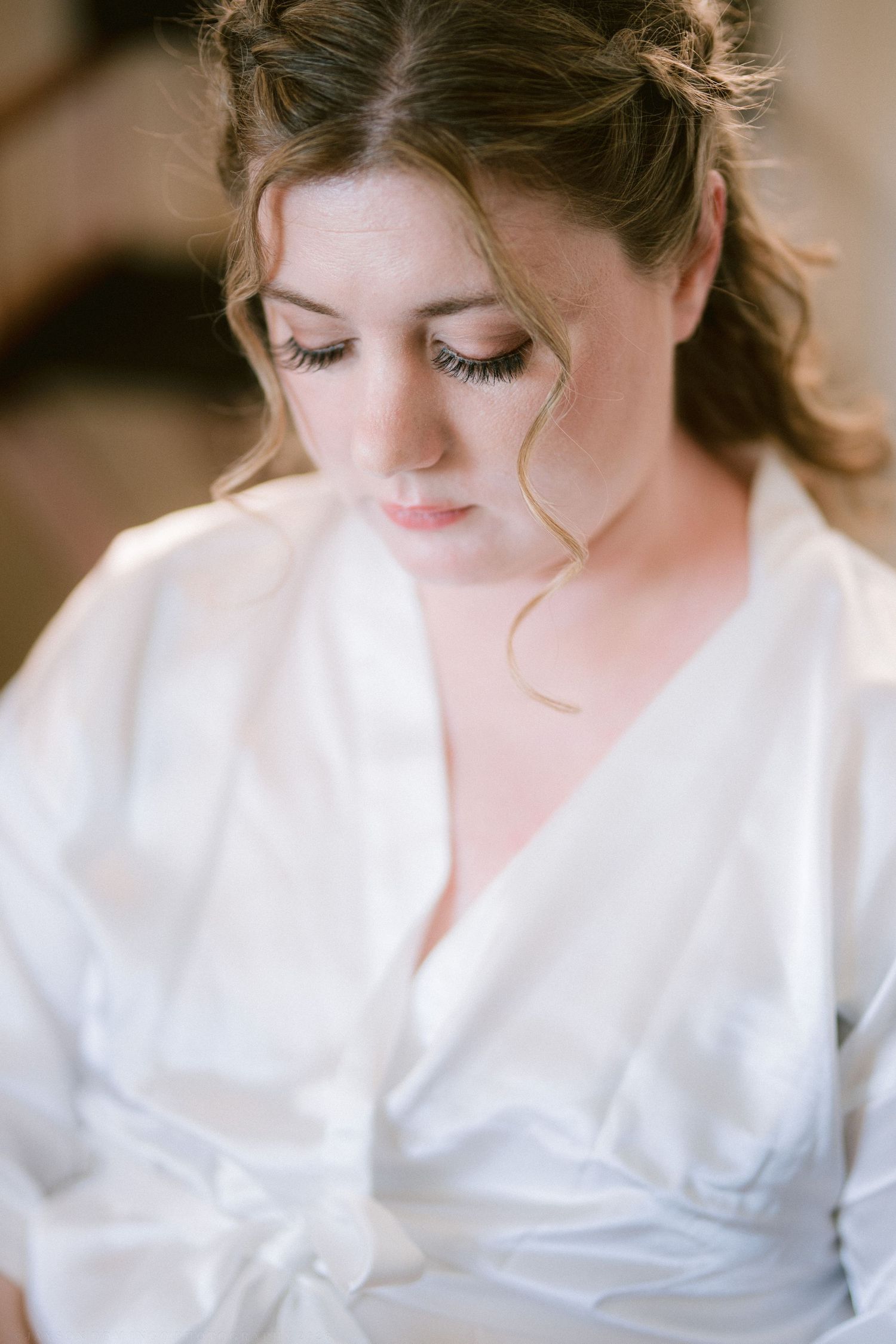 Bride in white satin robe with soft makeup and updo hairstyle looking down gracefully.