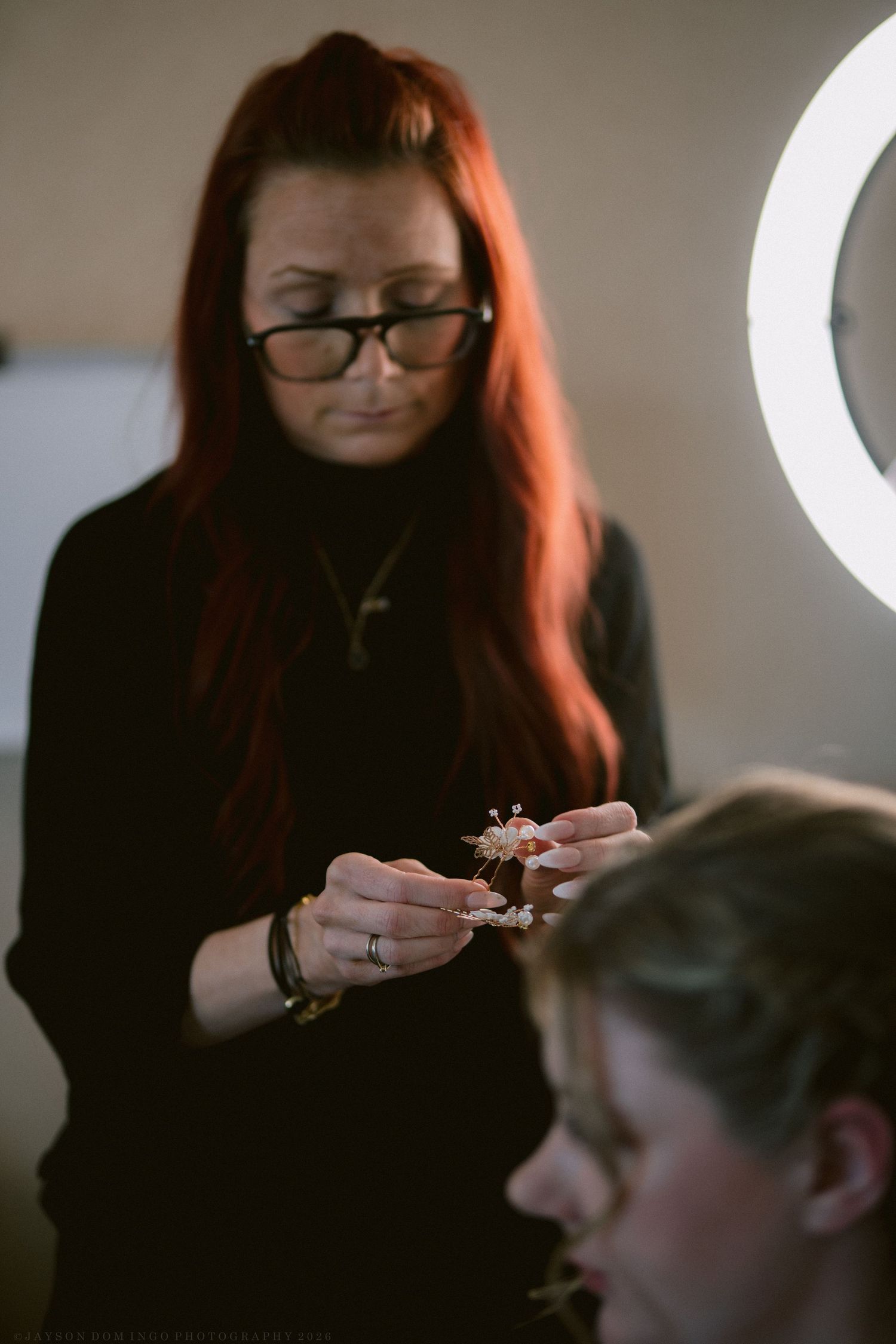 Red-haired hairstylist preparing floral hair accessory for bride under ring light during wedding prep.