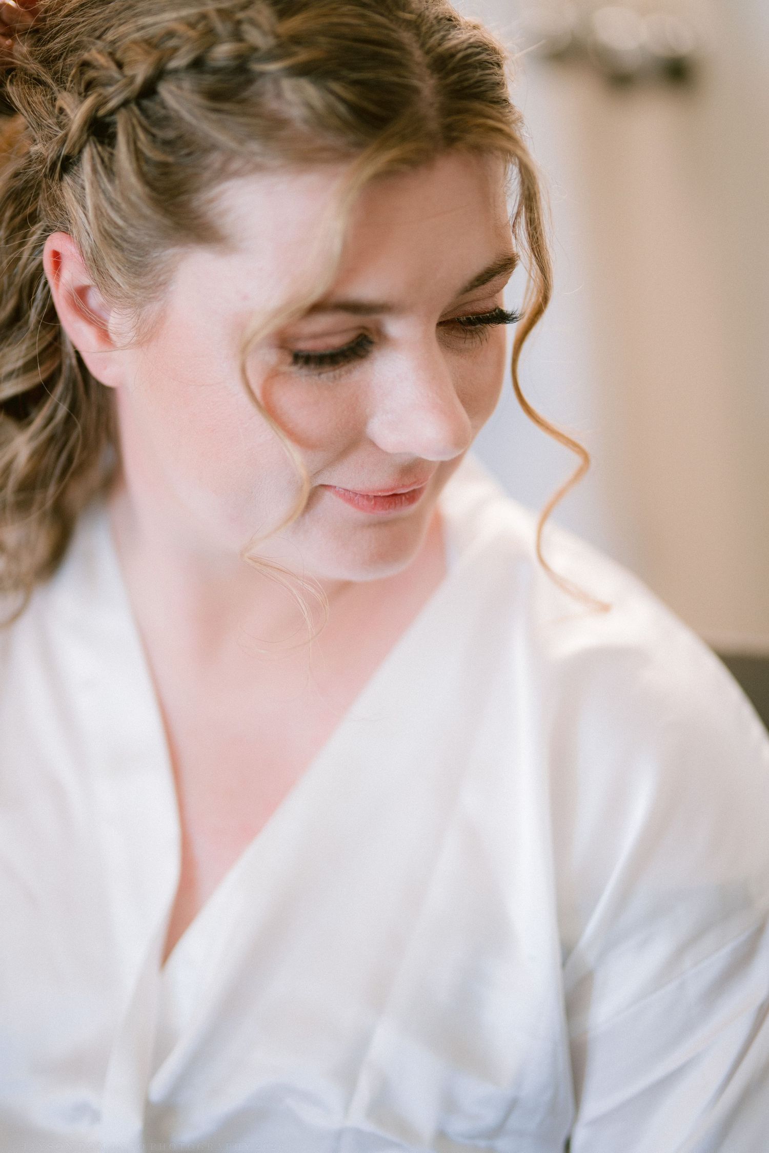 Beautiful bride in white satin robe with elegant braided updo and soft bridal makeup looking down.