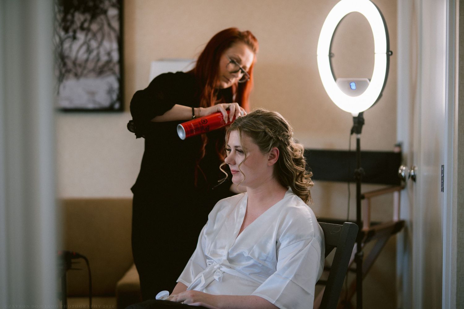 Smiling bride in white robe gets hair styled by red-haired stylist under ring light.