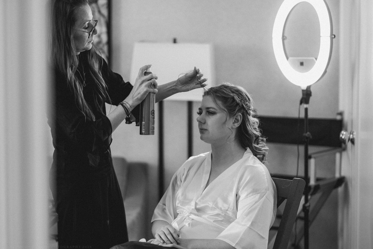Black and white photo of a bride in a white robe getting hair styled with hairspray before her wedding.