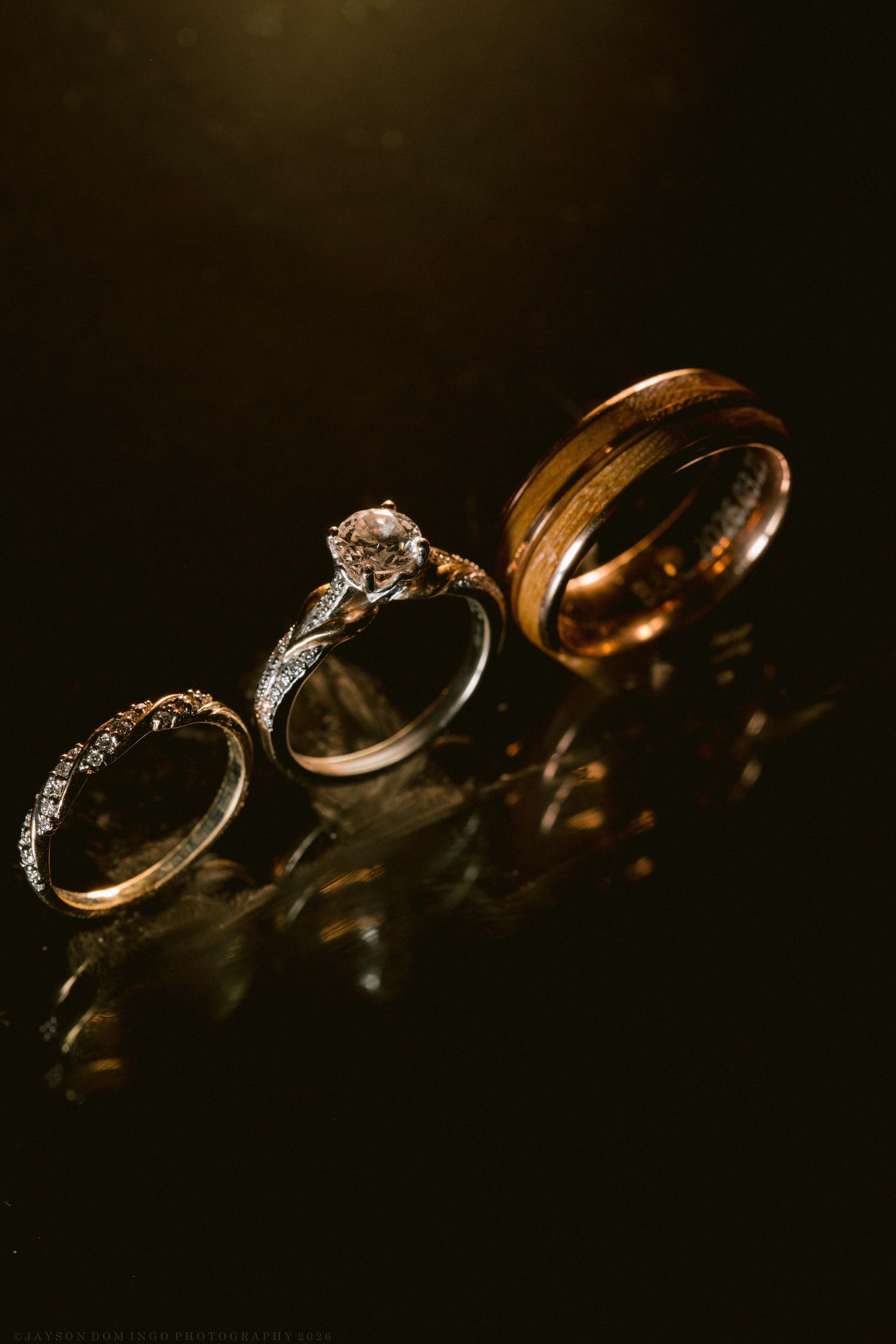 Three wedding rings displayed on a dark reflective surface, including a diamond engagement ring and a wooden band.