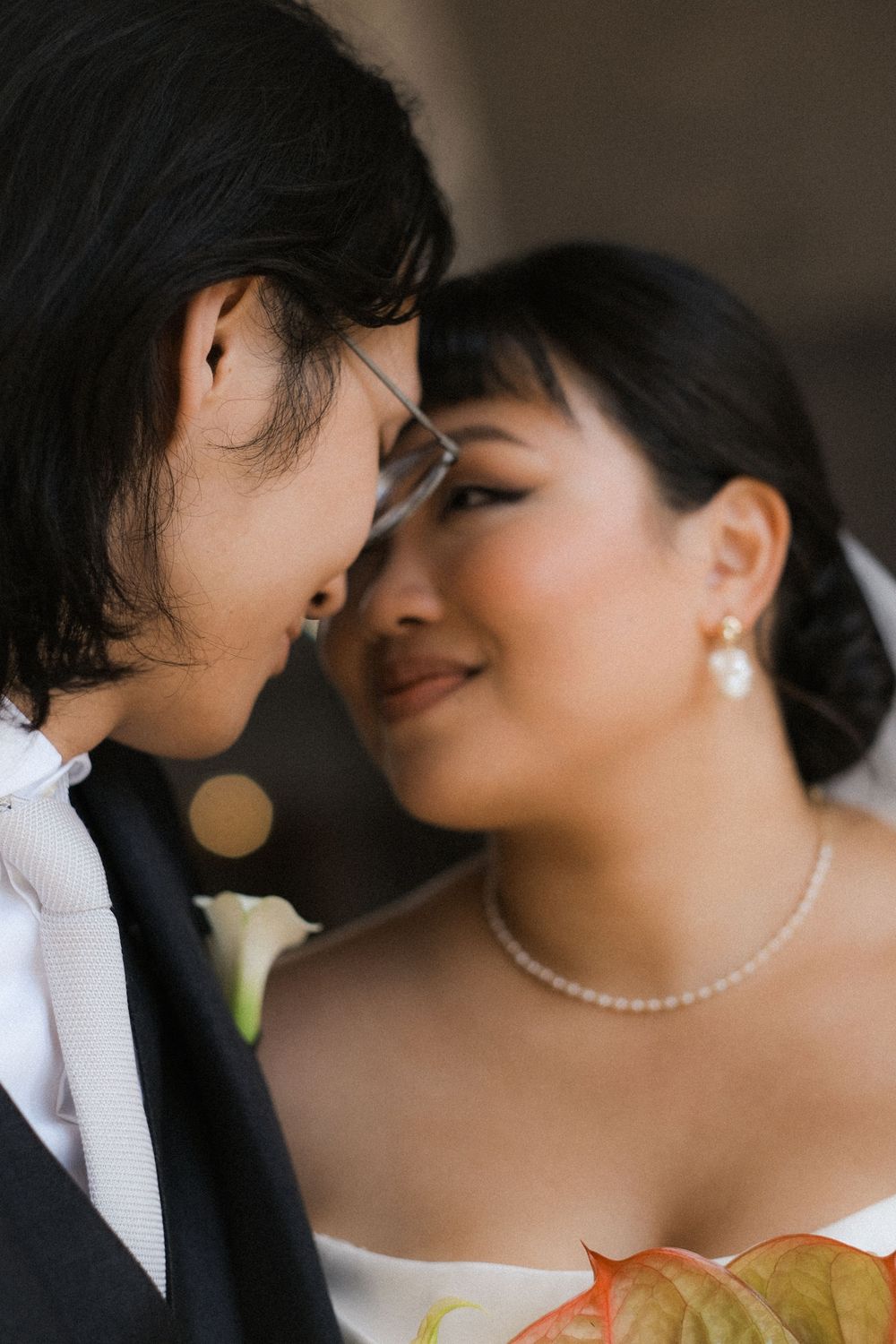 Asian bride and groom share an intimate forehead touch, bride wearing pearl jewelry and white dress on wedding day.